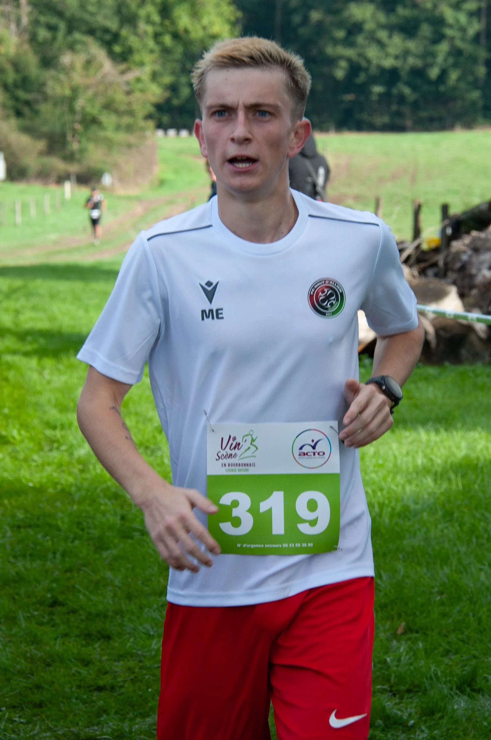 Un jeune homme court lors d'une course en plein air, portant un t-shirt blanc avec un logo et un dossard numéro 319, dans un paysage verdoyant avec des arbres en arrière-plan.