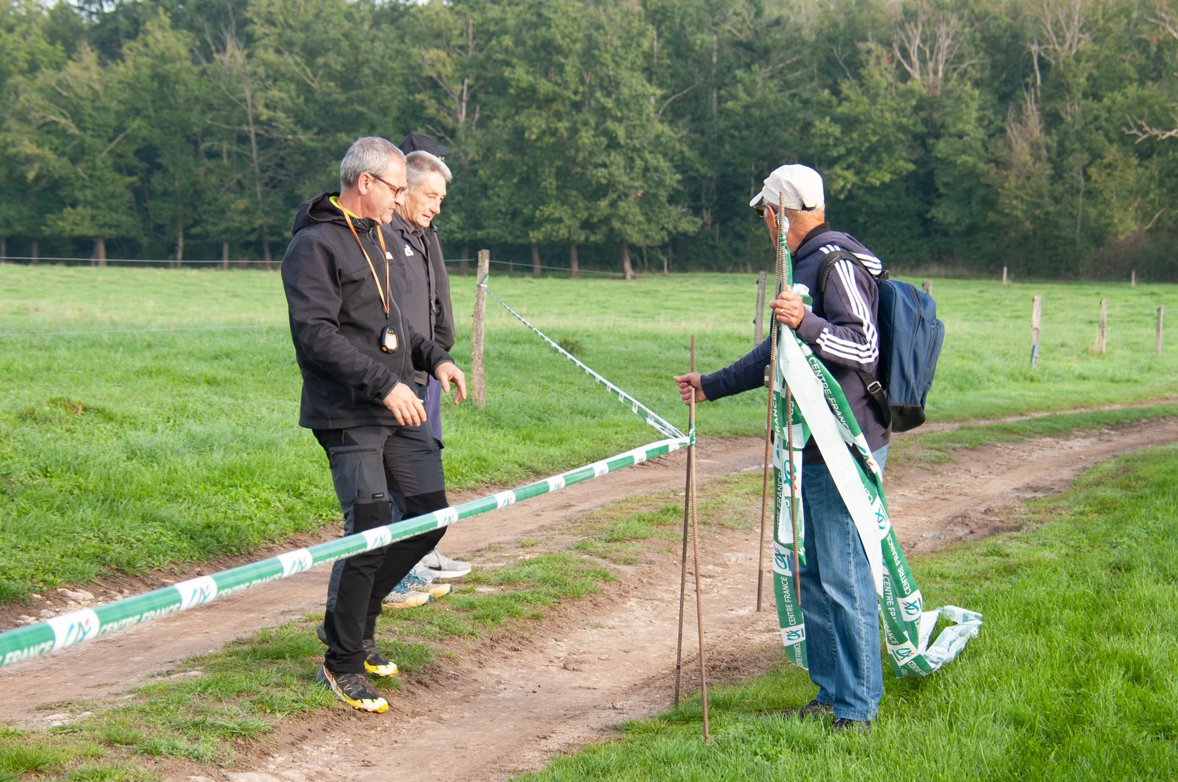 Trois hommes discutent à côté d'une corde délimitant un sentier dans un champ vert avec des arbres en arrière-plan. Deux hommes portent des vêtements de sport noirs, un porte une casquette blanche et un sac à dos, ils semblent planifier ou examiner l