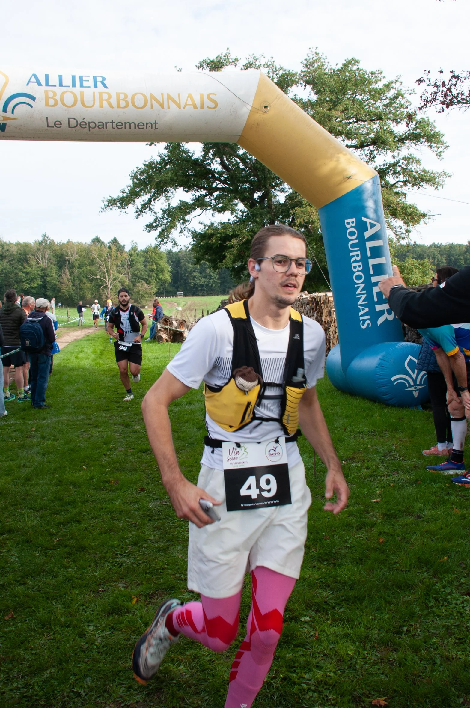 Un homme court lors d'une course en plein air, portant un dossard avec le numéro 49, un t-shirt blanc, un short beige, des collants roses, et une petite gourde, sous une arche de départ ou d'arrivée portant l'inscription "ALLIER BOURBONNAIS".