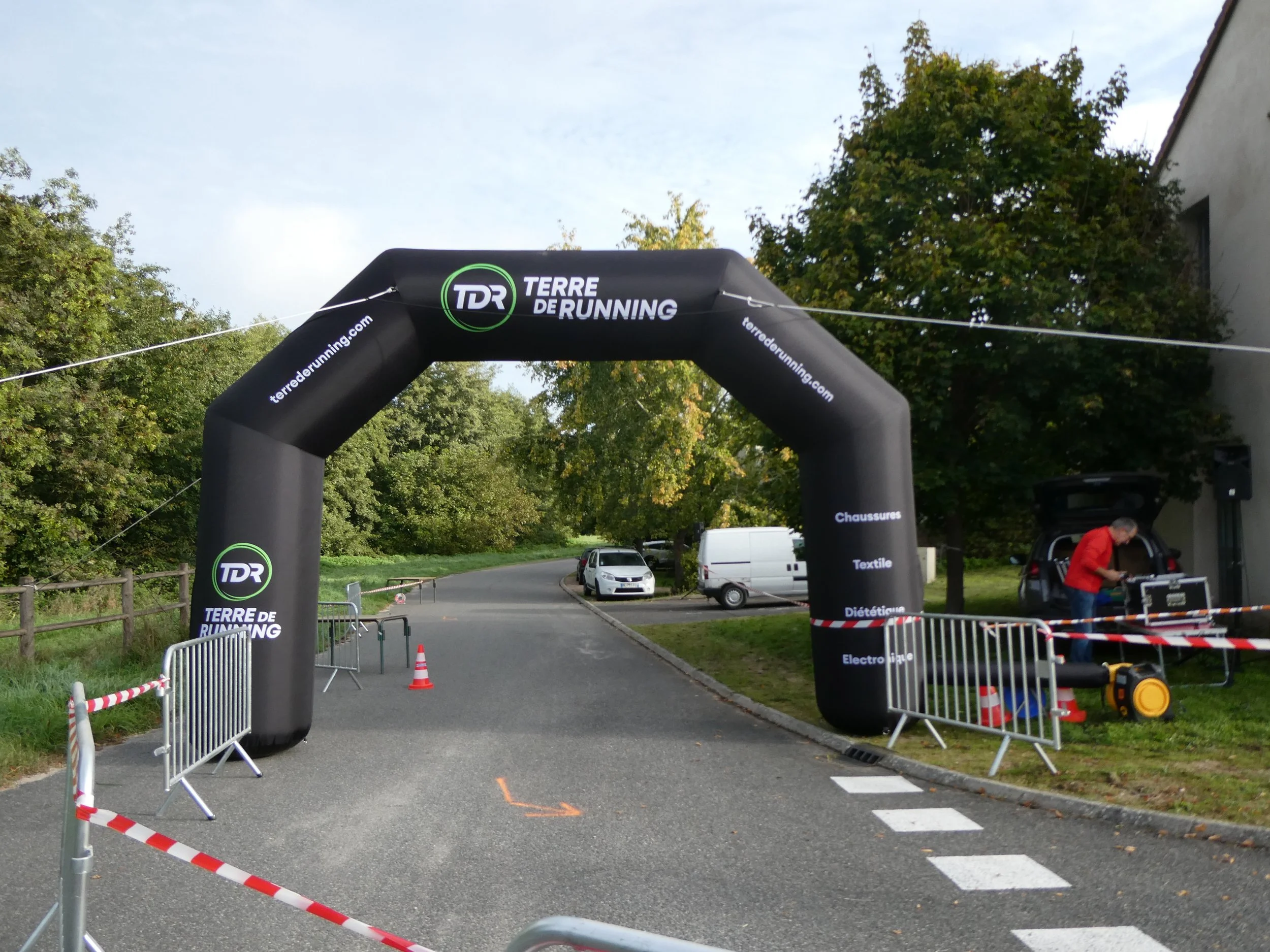 Un arc gonflable publicitaire pour un événement de course, avec des barrières et des véhicules en arrière-plan, située sur une route bordée d'arbres.