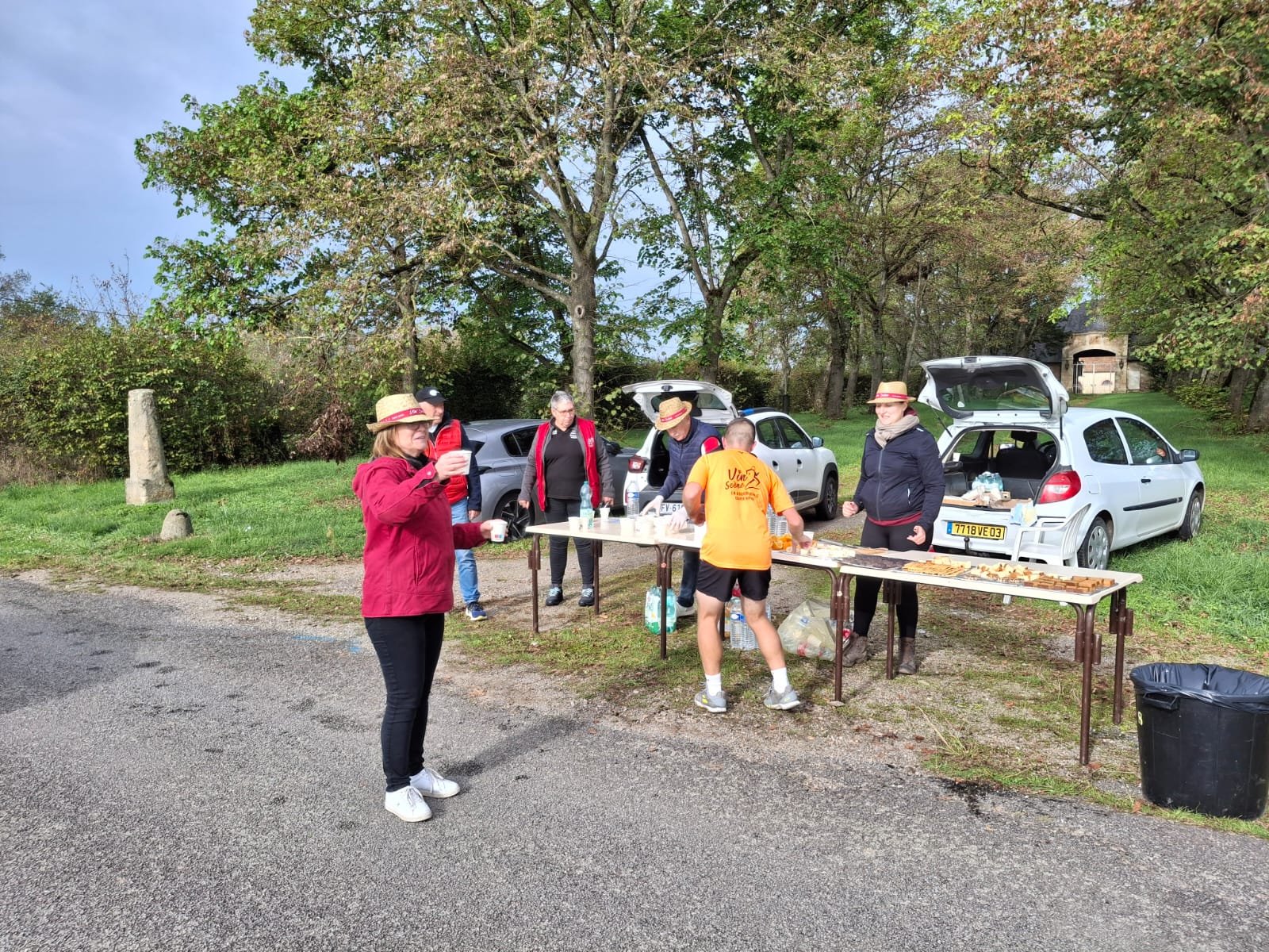 Groupe de personnes faisant une pause lors d'un événement en plein air, avec un stand de nourriture et des voitures garées sous les arbres.