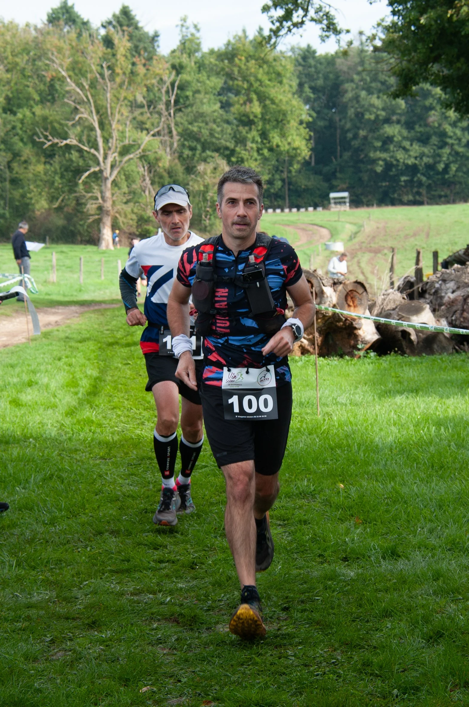 Deux hommes en course à pied dans un parc vert avec arbres et herbe, portant des vêtements de sport, numéros de course 100 et 1.