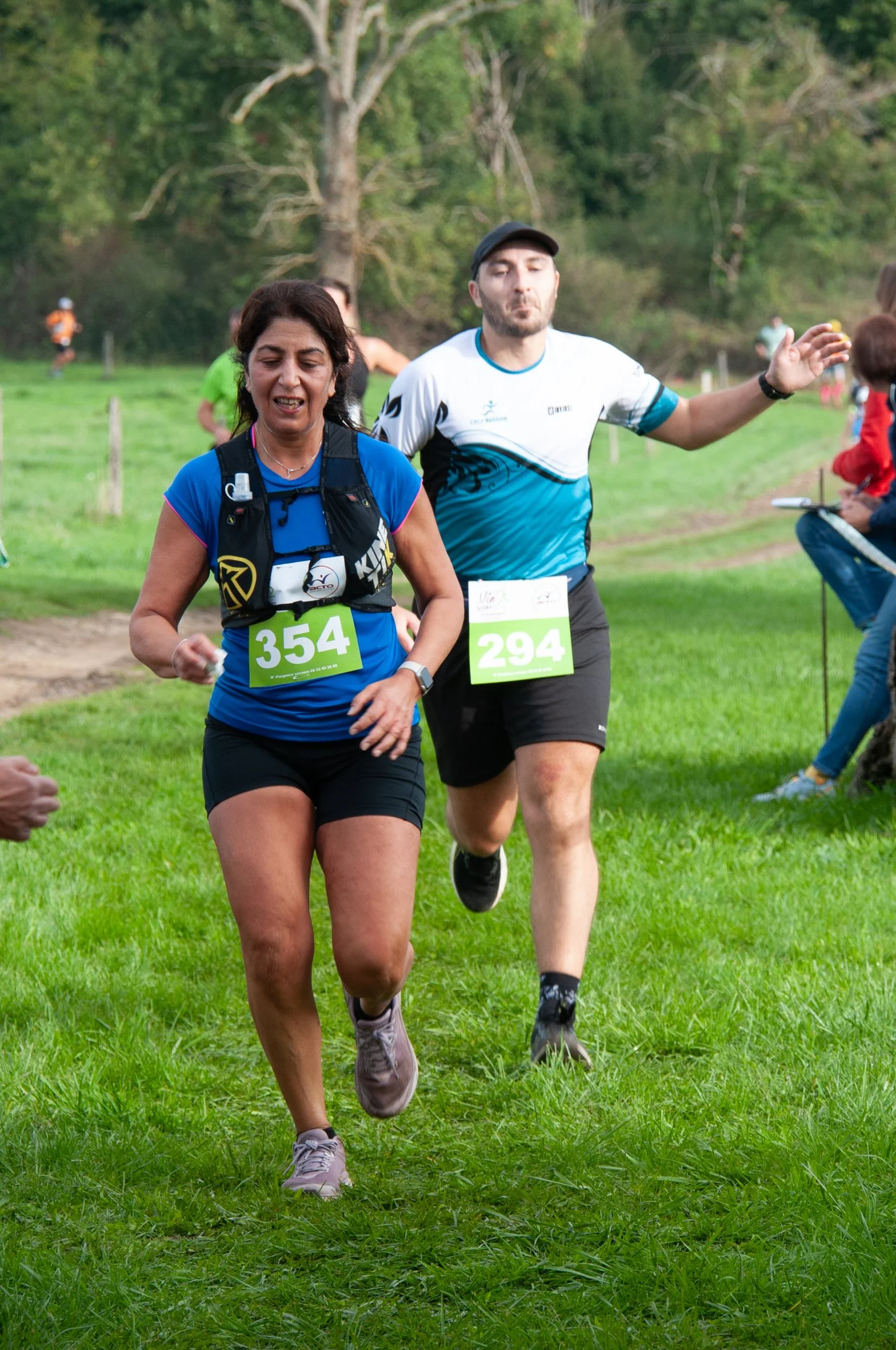 Deux coureurs participant à une course en extérieur, portant des dossards verts avec les numéros 354 et 294, courant sur un chemin herbeux avec des arbres en arrière-plan.