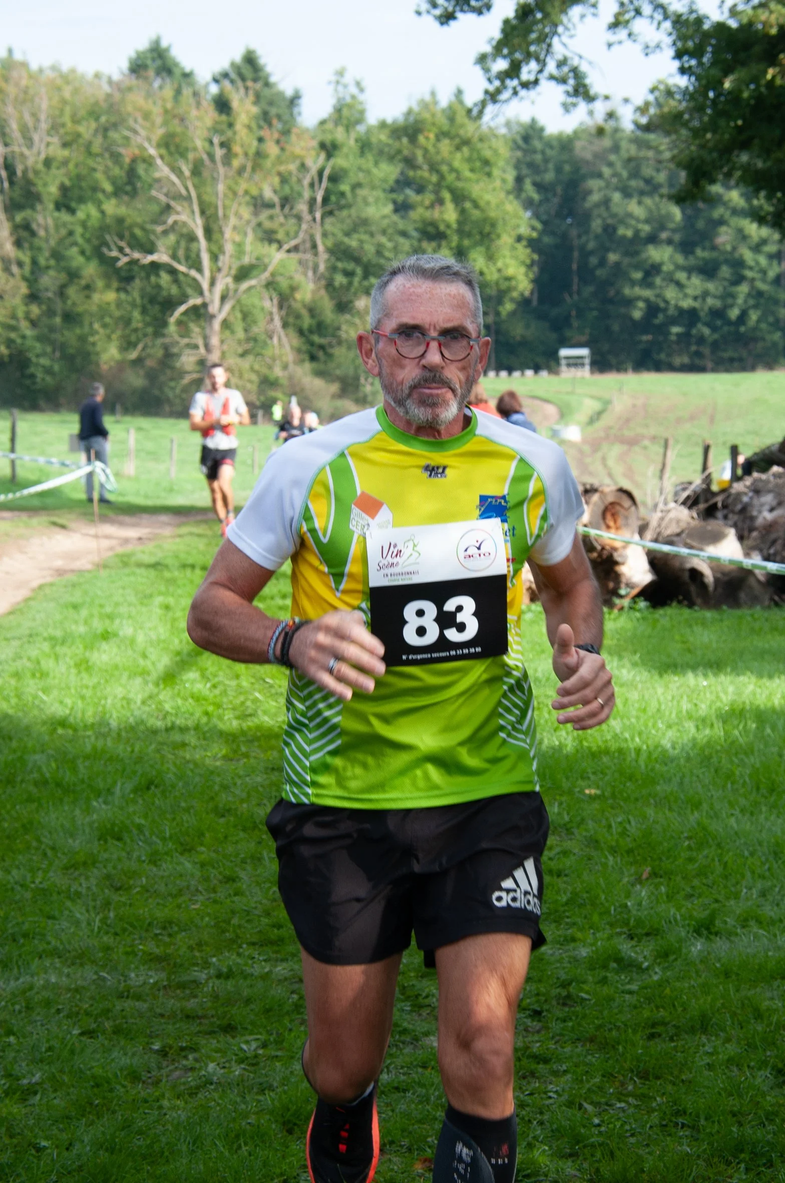 Un homme participe à une course en extérieur, portant un dossard numéro 83, vêtu d'une chemise de course verte et blanche, avec des arbres en arrière-plan.