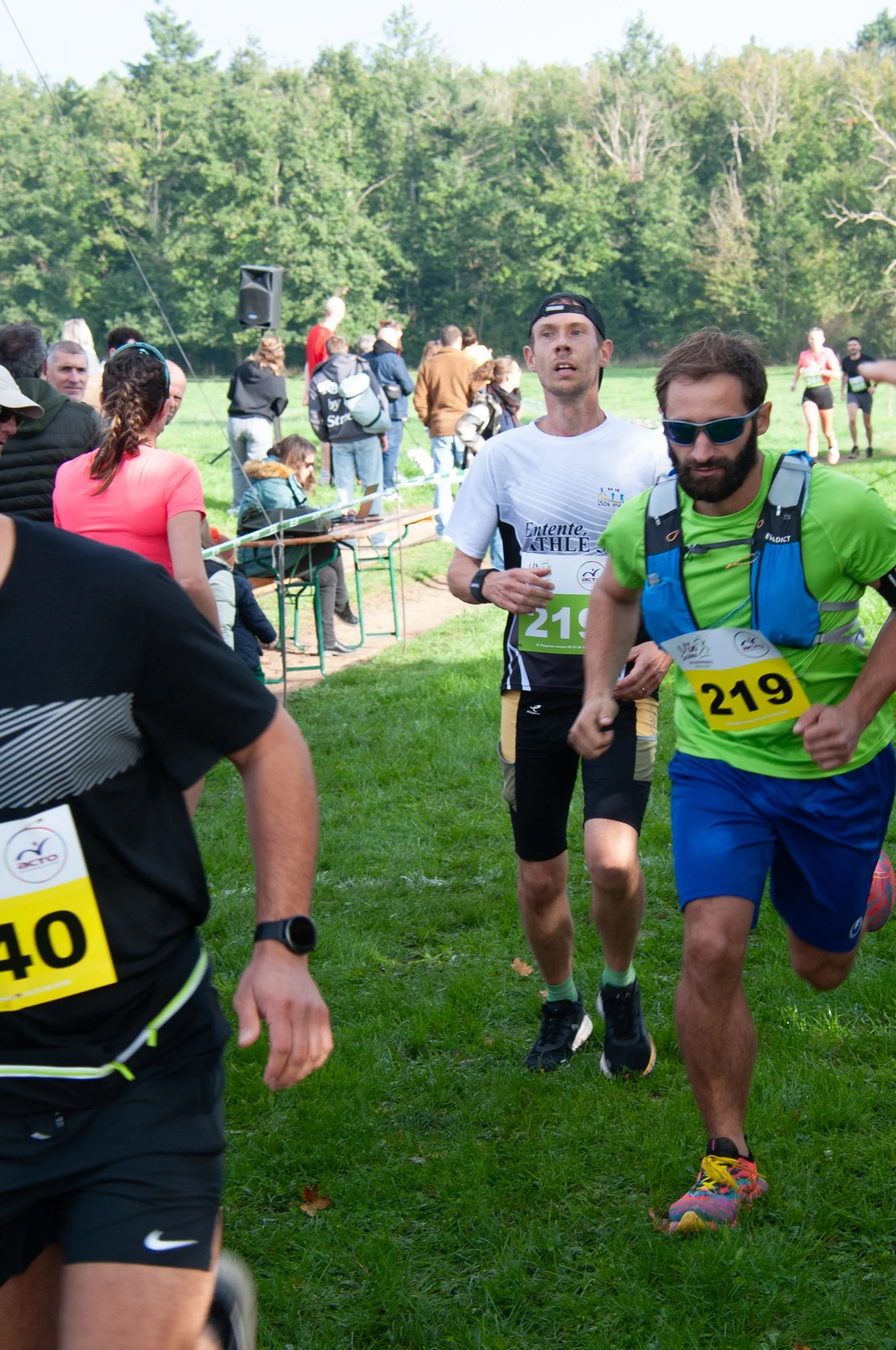 Les coureurs participent à une course en extérieur, avec des spectateurs et des assistants autour dans un environnement naturel verdoyant.