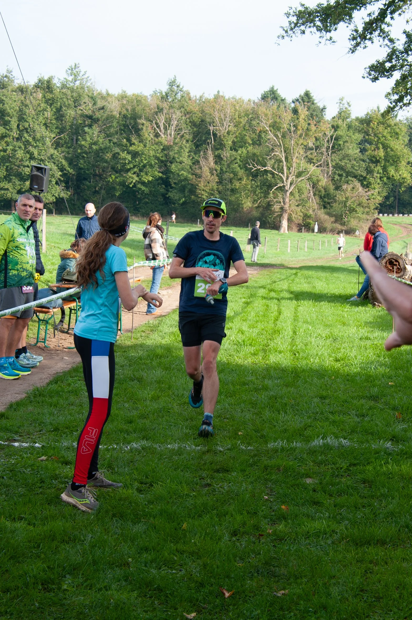 Un homme court vers la ligne d'arrivée lors d'une course à pied en plein air, avec des spectateurs à ses côtés et des arbres dans le fond.
