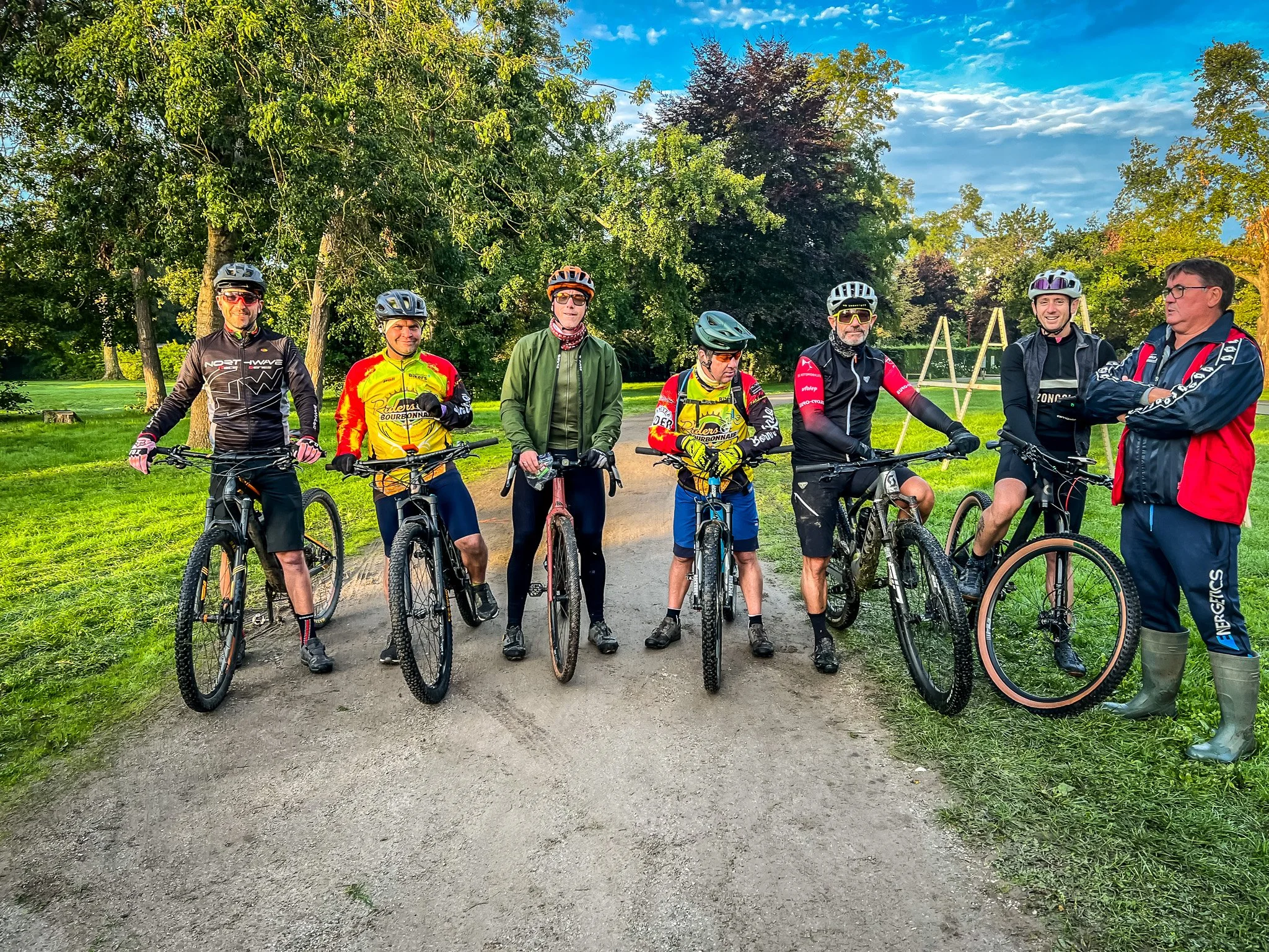 Groupe de sept personnes en tenue de cyclisme, avec des casques, posant avec leurs vélos dans un parc verdoyant par une journée ensoleillée.