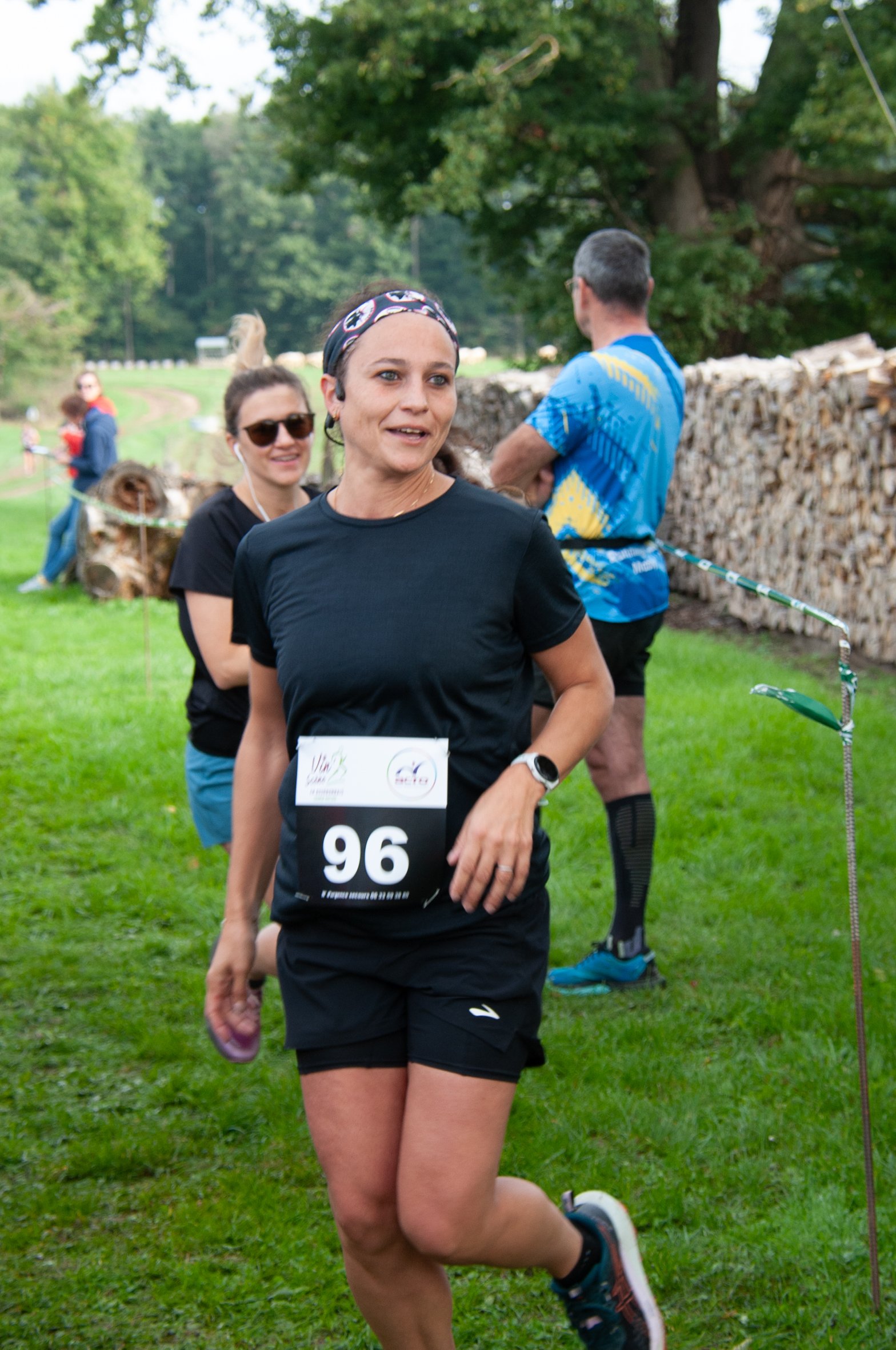 Femme courant lors d'une compétition, portant un numéro 96, un t-shirt noir, des shorts noirs, une montre connectée, et un bandeau sur la tête, avec d'autres participants en arrière-plan dans un parc avec des arbres.