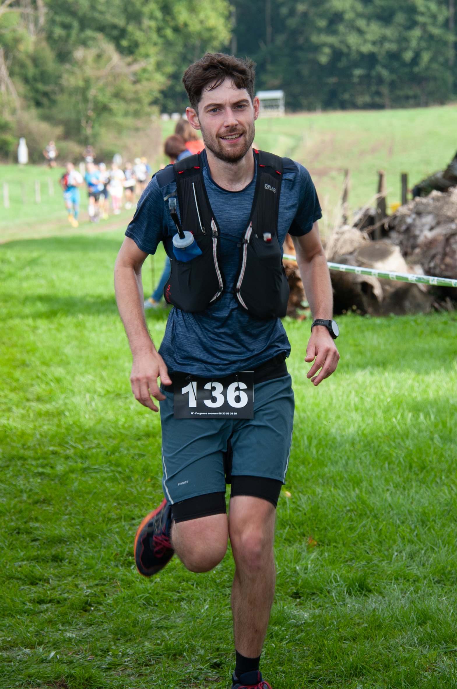 Un homme en tenue de course, portant un dossard numéro 136, participe à une course en plein air, avec un fond de verdure et d'autres coureurs en arrière-plan.