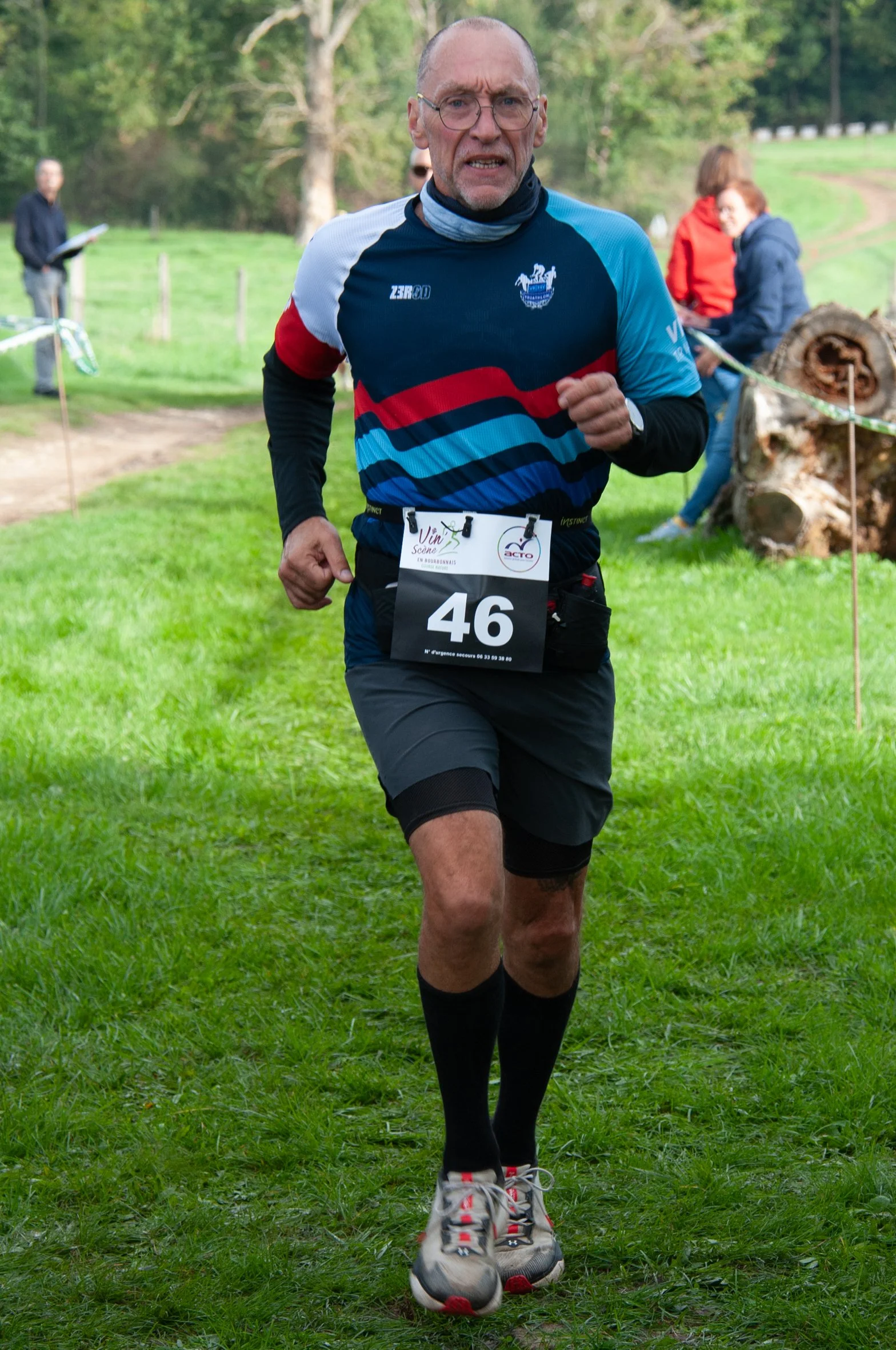 Un homme en course lors d'une compétition en plein air, portant un maillot bleu avec des motifs rouges et bleus, un numéro 46 sur le ventre, et des chaussures de course dans un espace boisé verdoyant.