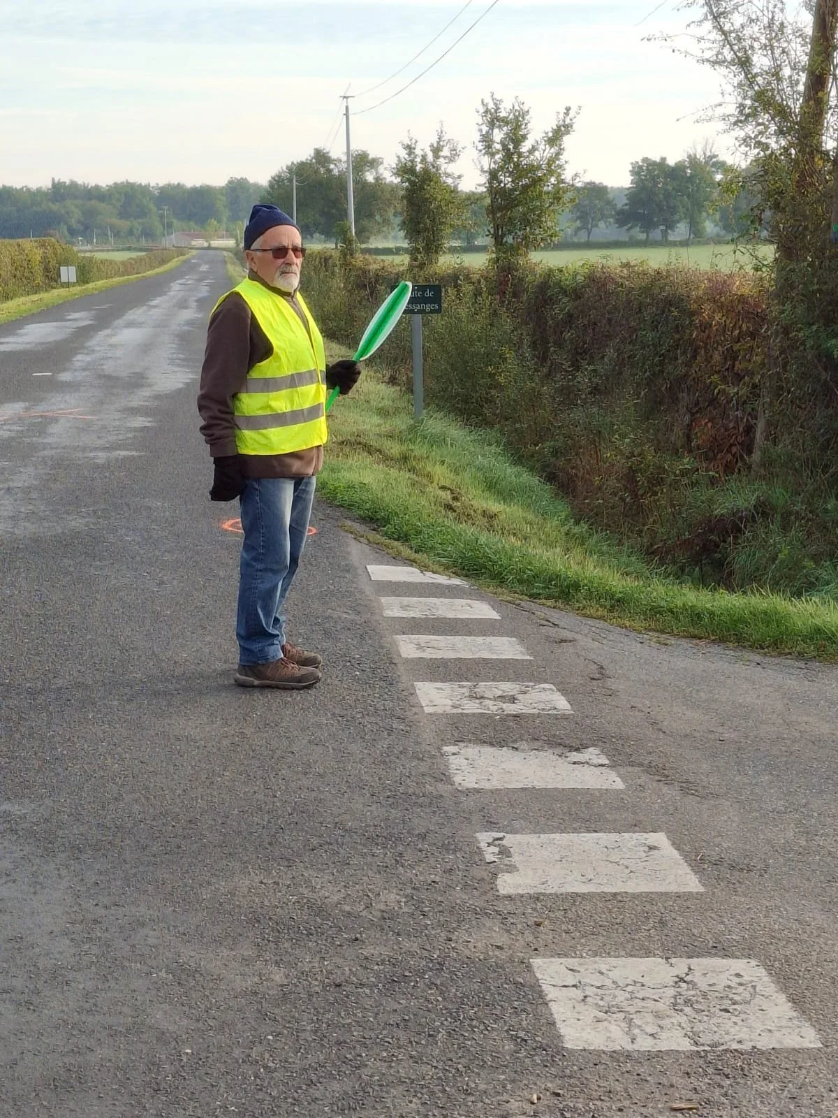 Un homme avec un gilet jaune et des lunettes, tenant un ballon en plastique vert, se tient sur le bord d'une route de campagne avec des champs et des arbres en arrière-plan.