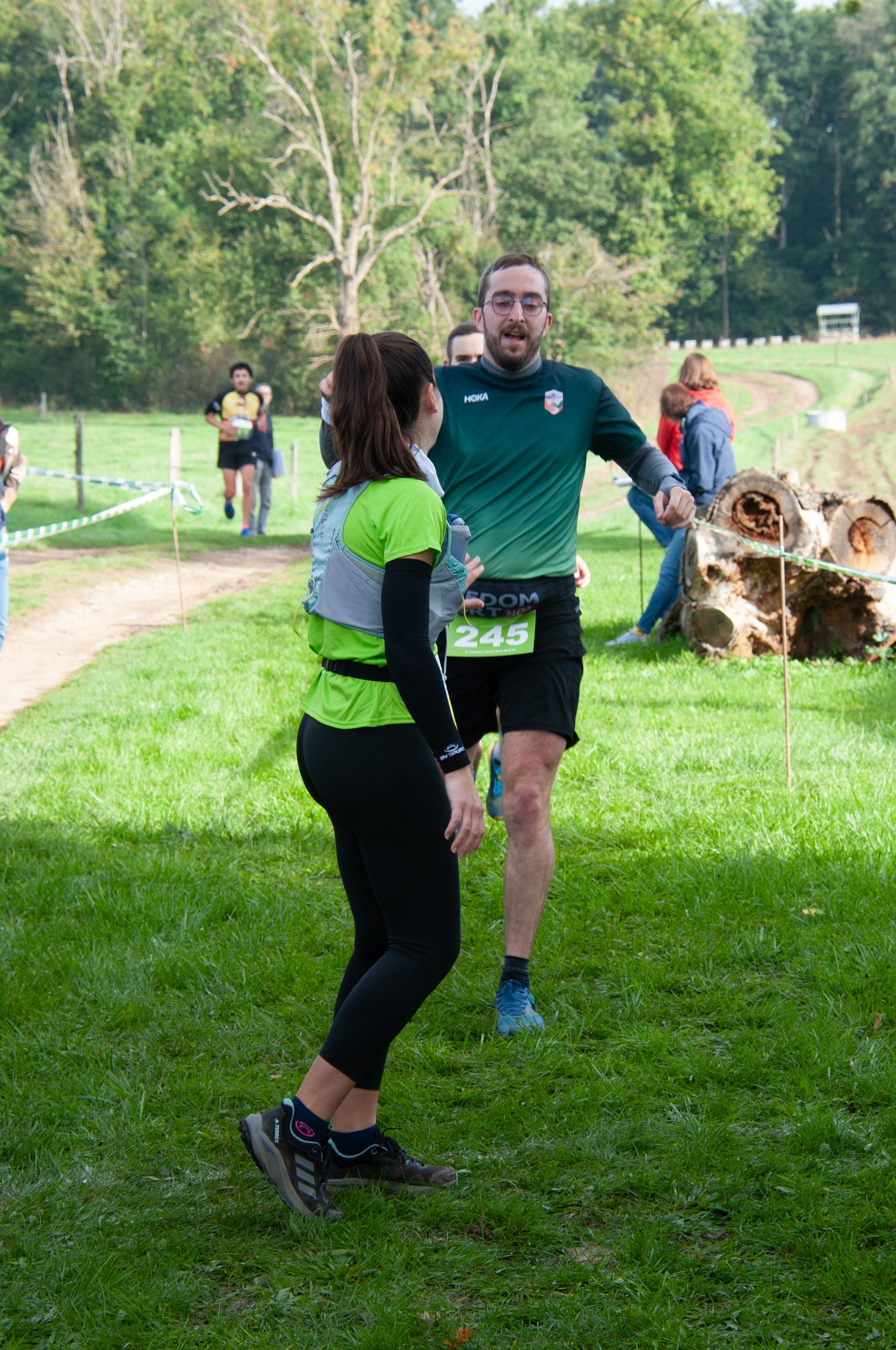 Un homme et une femme discutent après une course en extérieur, dans un cadre naturel avec des arbres et de l'herbe, pendant qu'ils se regardent et que d'autres coureurs se trouvent en arrière-plan.
