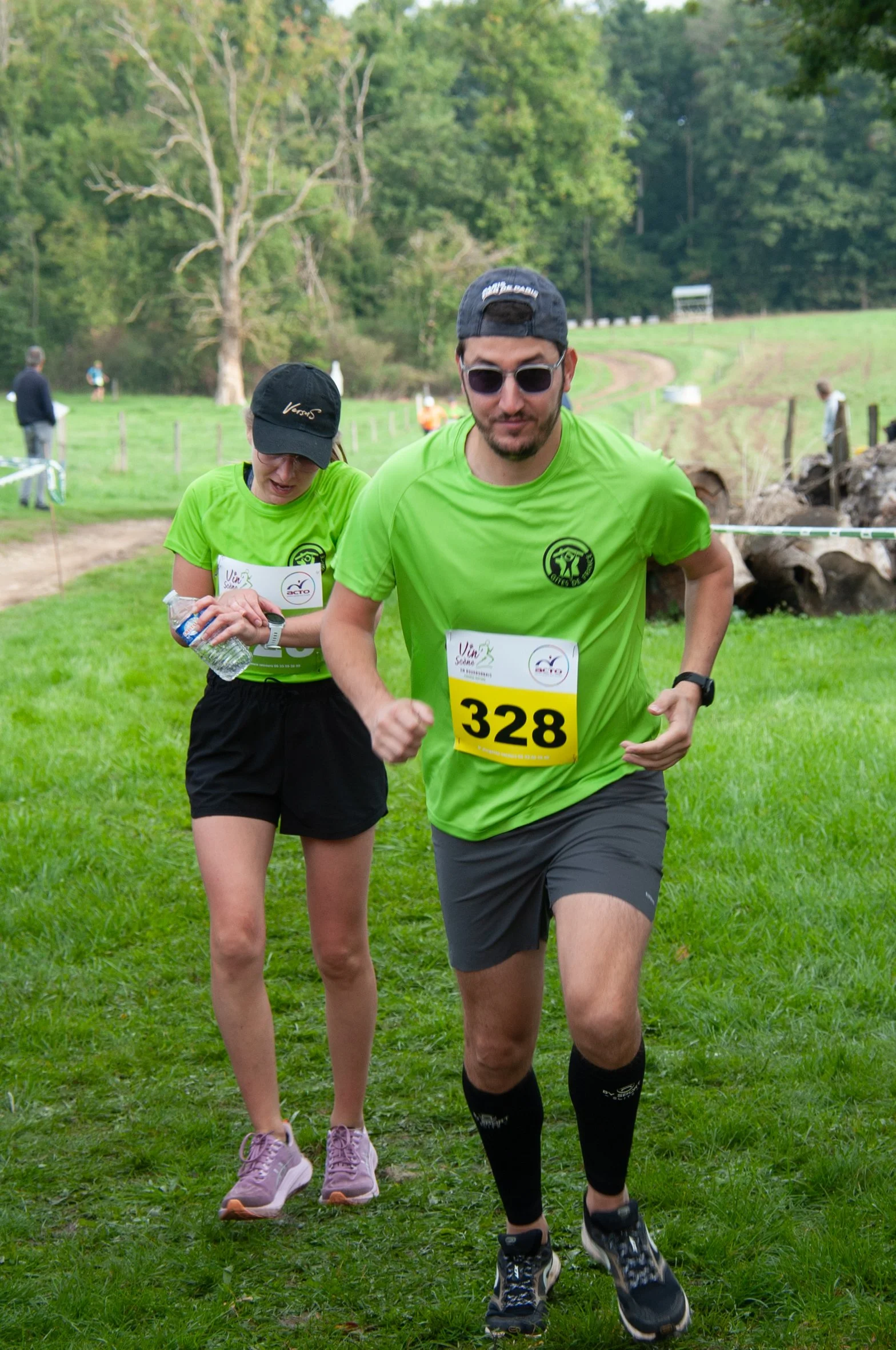 Deux coureurs en t-shirts verts participent à une course dans la nature, avec un homme en avant portant le numéro 328 et une femme derrière lui, tous deux portant des casquettes et des vêtements de sport, dans un environnement verdoyant.