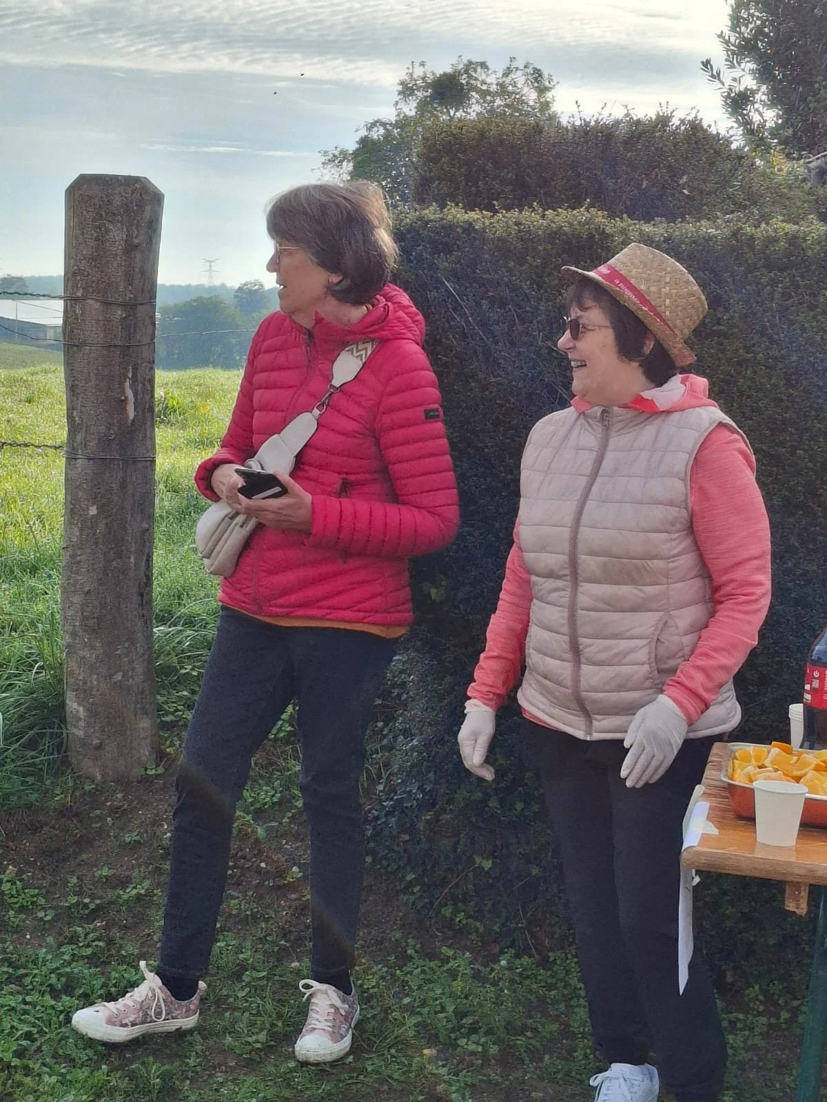 Deux femmes souriantes en vêtements décontractés, debout dehors près d'une table avec des boissons et des morceaux d'orange, en train de discuter.