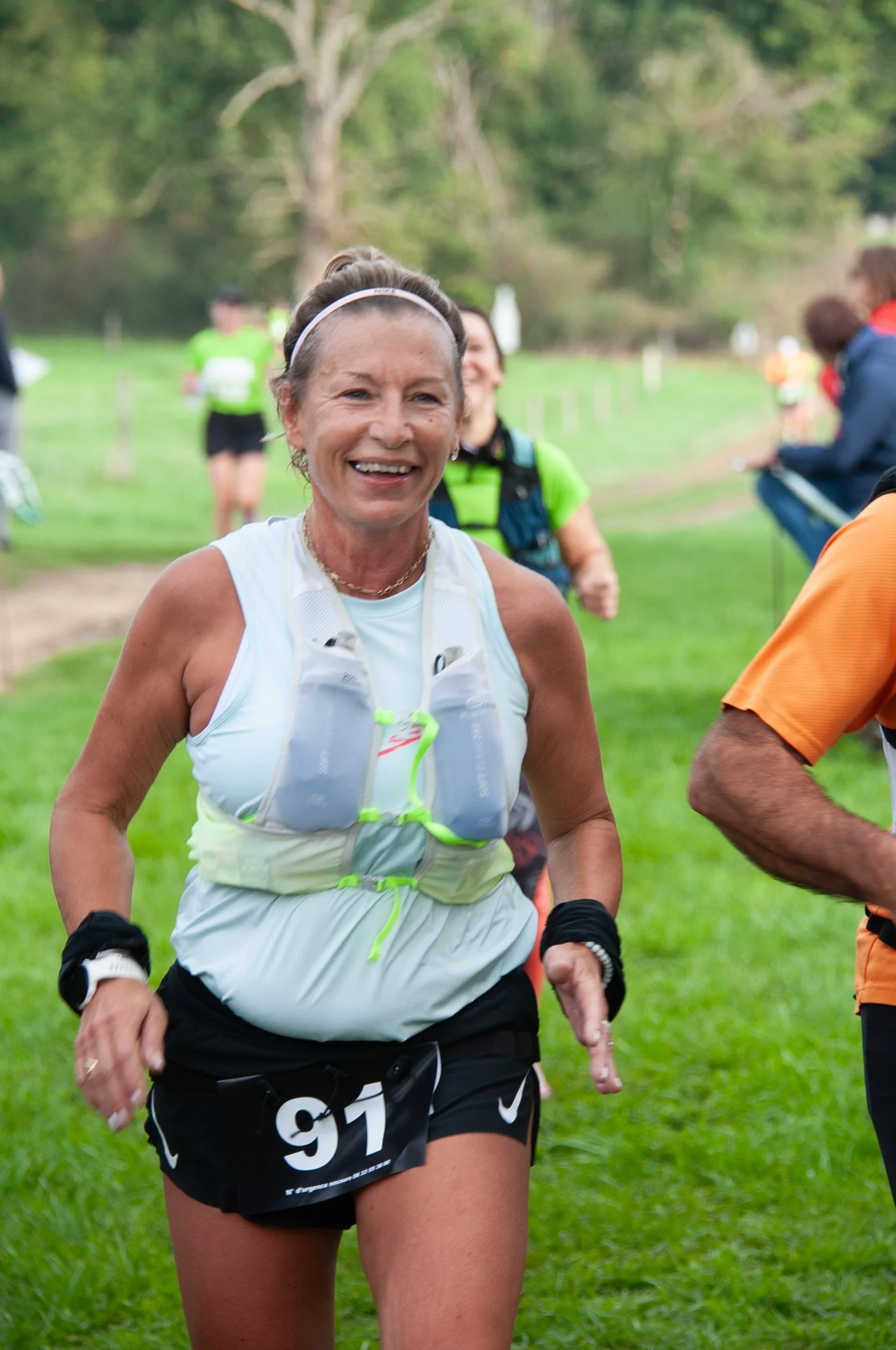 Femme courant lors d'une course en plein air, portant un dossard numéro 97, vêtue de vêtements de sport, avec un sourire et l'arrière-plan d’un parc ou d’un sentier naturel.