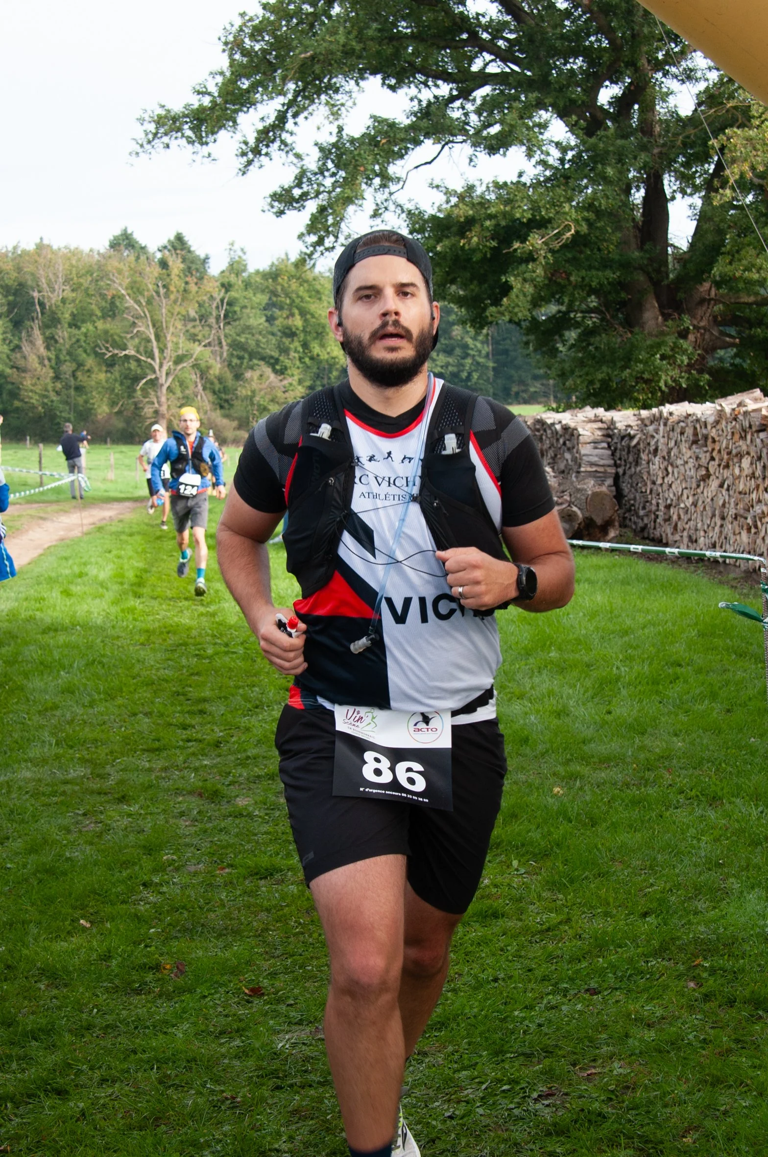 Un homme participe à une course en plein air, portant un t-shirt de sport, un numéro de course 86, une montre et un sac à dos, avec d'autres coureurs en arrière-plan dans un paysage vert et boisé.