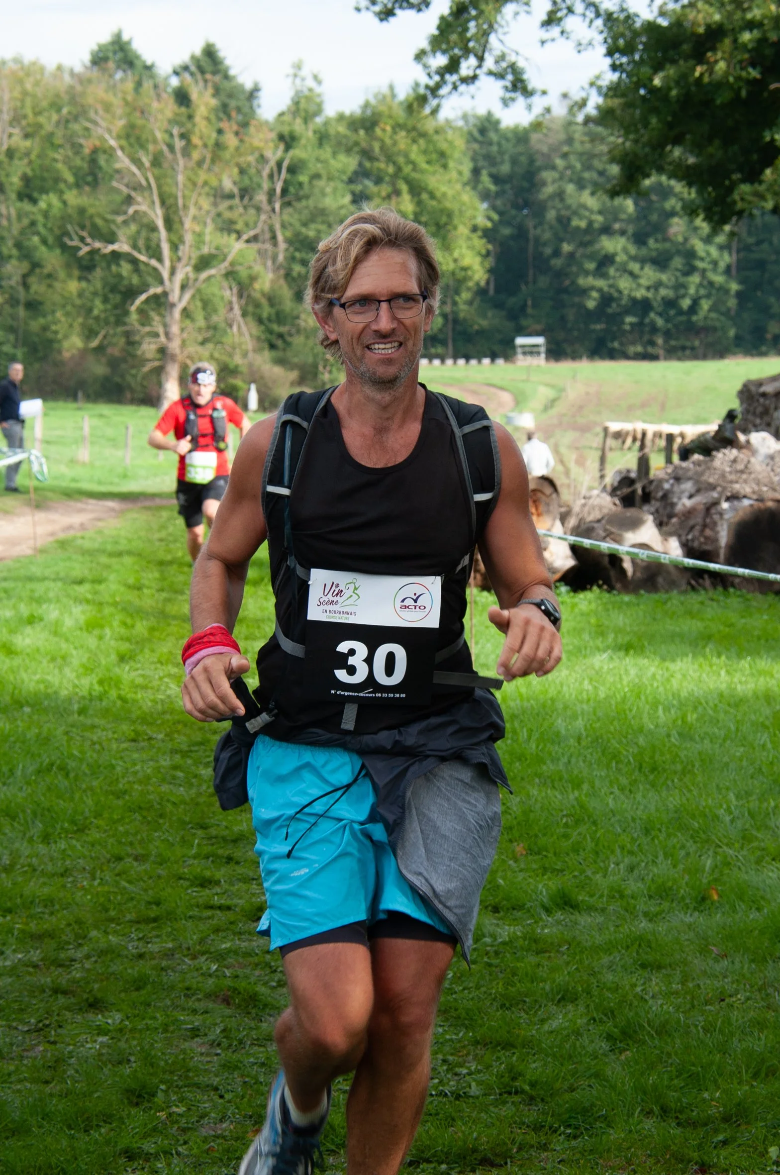 Un homme courrant lors d'une course en plein air, portant un t-shirt noir, un short bleu et gris, avec un numéro de course 30, dans un environnement naturel avec des arbres et de l'herbe.