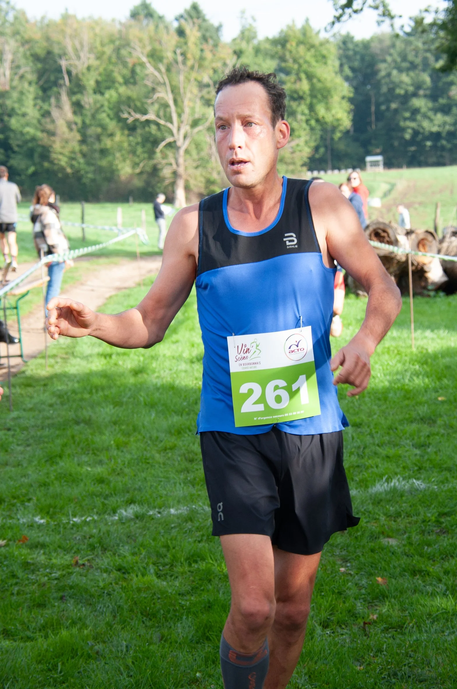 Un homme court lors d'une course en plein air, portant un dossard numéro 261. Il porte un débardeur bleu et noir et un short noir, avec un arrière-plan de verdure et quelques spectateurs.