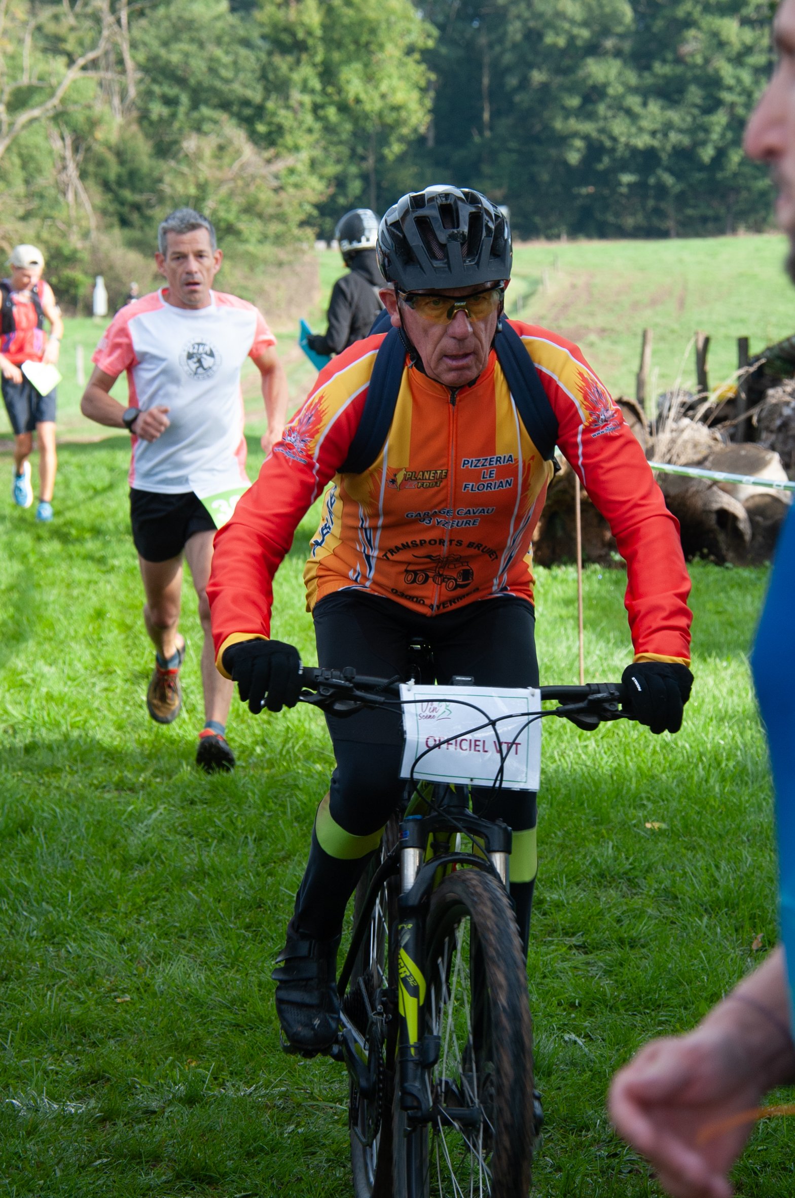 Un homme portant un casque et des lunettes de soleil fait du vélo lors d'une course en plein air, avec d'autres coureurs à pied derrière lui sur un chemin herbeux entouré d'arbres.