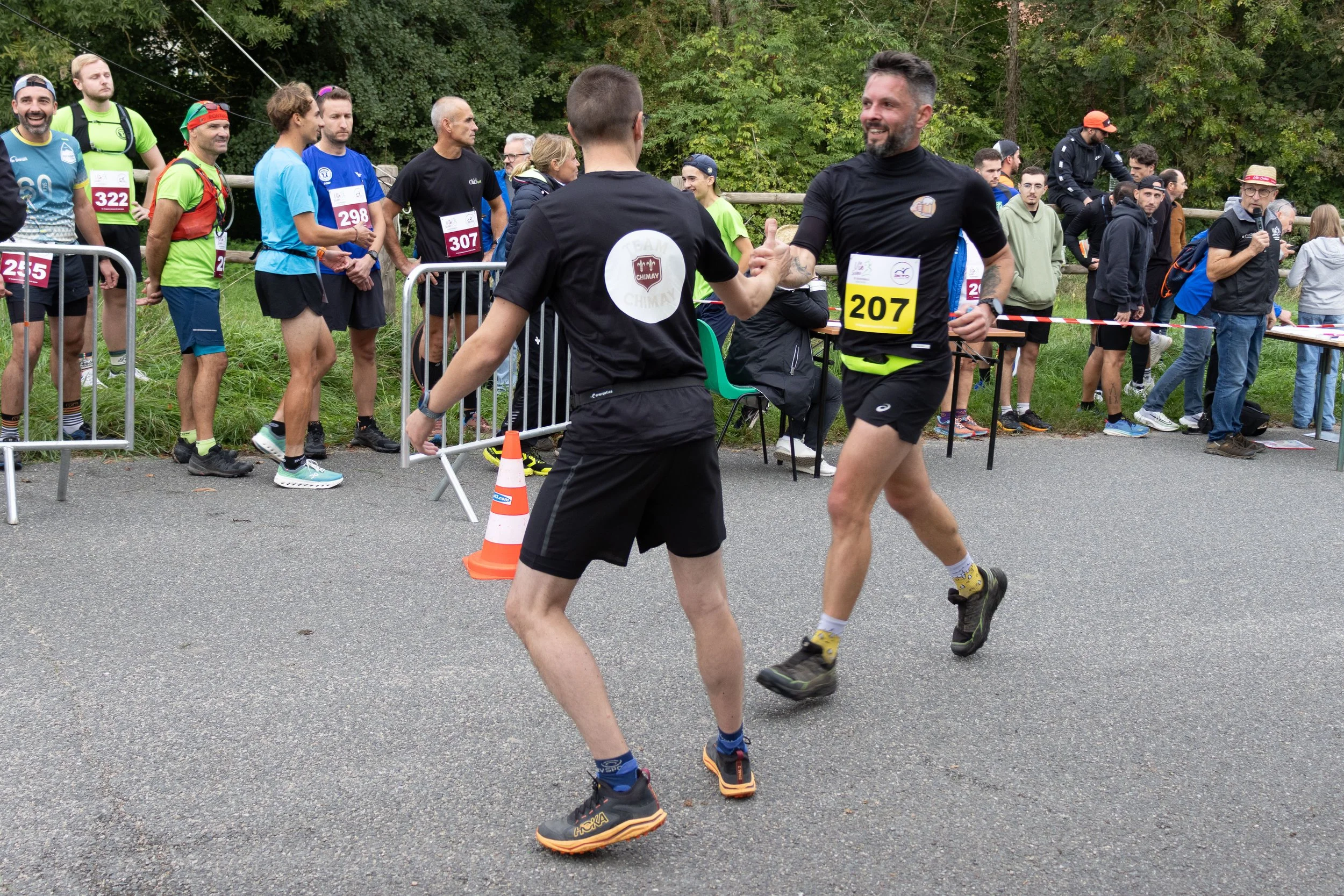 Deux coureurs de marathon se saluent ou échangent une fièvre à la fin de la course, entourés d'autres participants et spectateurs, avec un espace vert en arrière-plan.