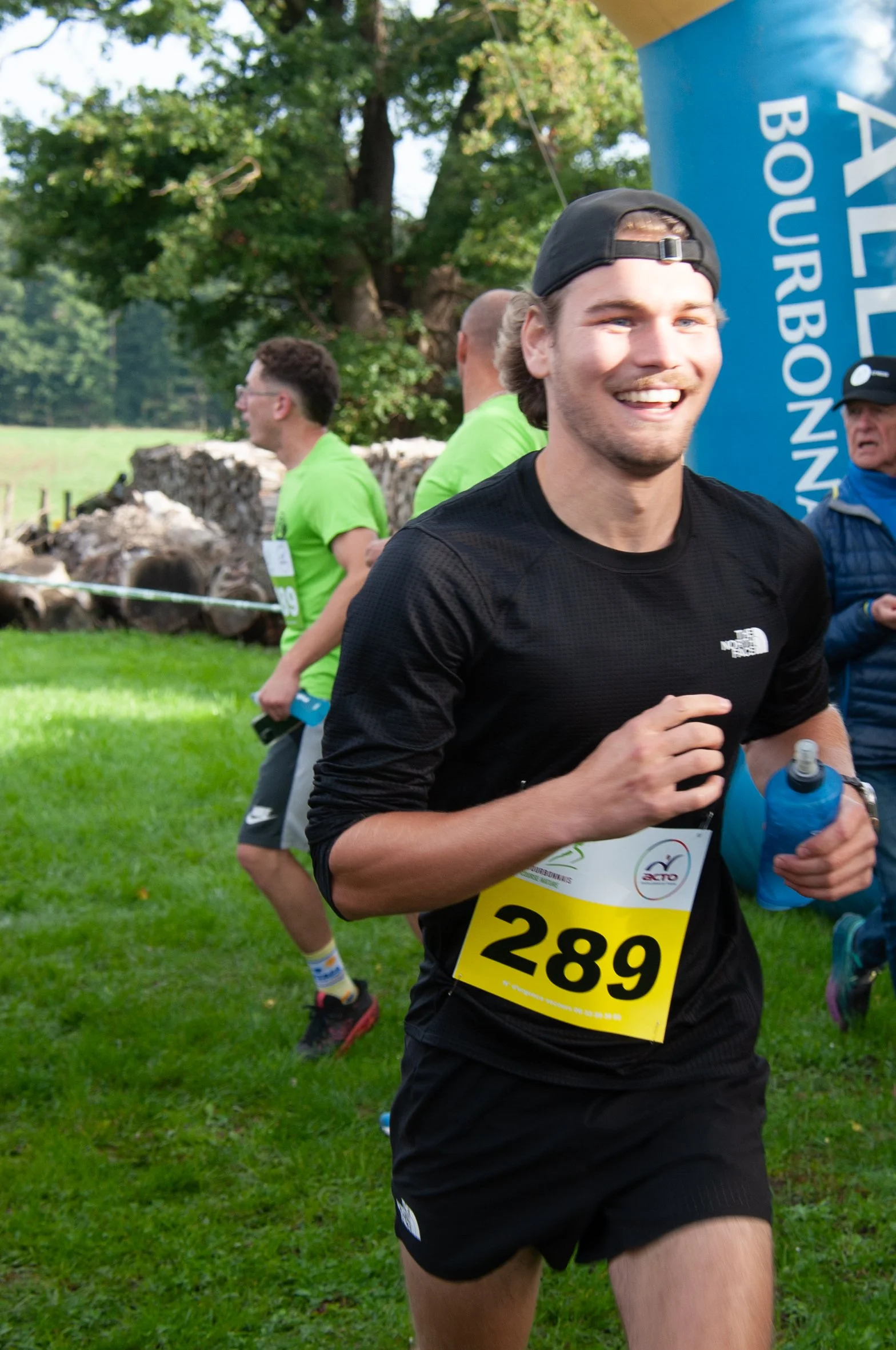 Un jeune homme souriant qui participe à une course en plein air, portant un numéro 289, vêtu d'un t-shirt noir, avec des arbres en arrière-plan.