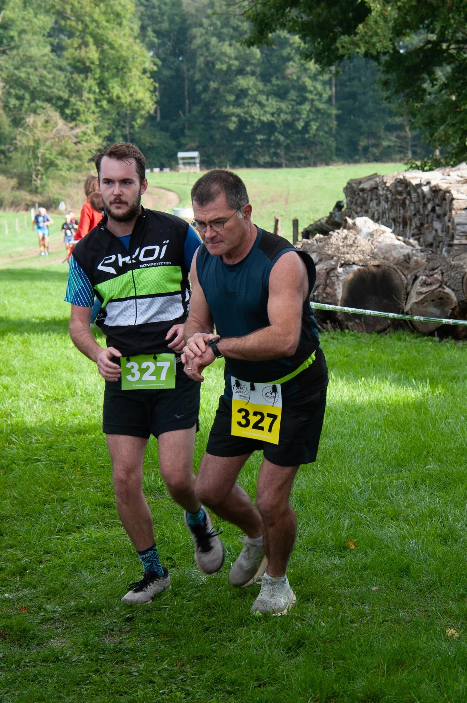 Deux hommes participent à une course en plein air, portant des numéros 327. L'un d'eux regarde sa montre tandis que l'autre le soutient. En arrière-plan, d'autres coureurs avancent dans un environnement verdoyant avec des arbres et des rondins de boi