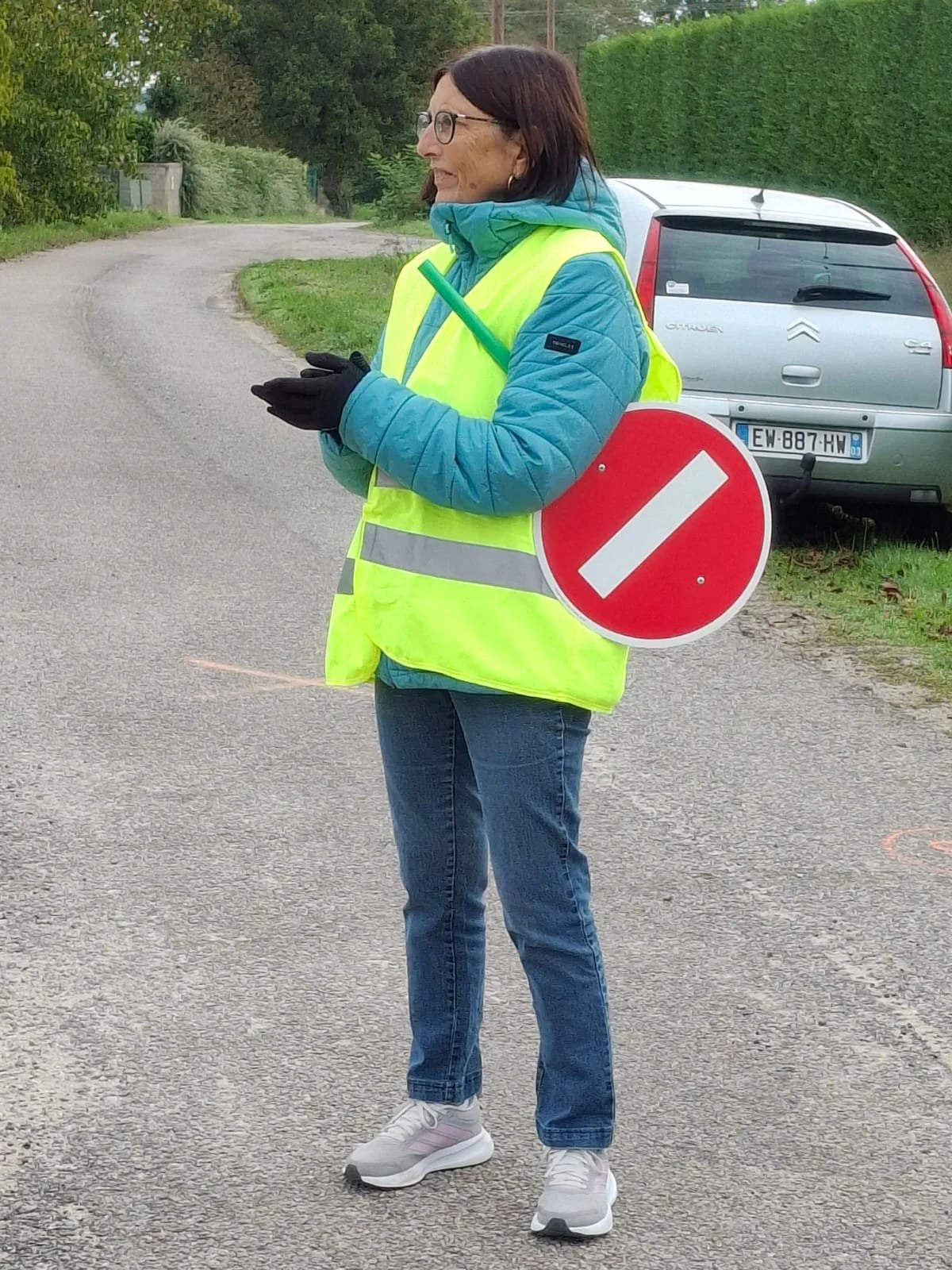 Femme portant un gilet jaune de signalisation avec un panneau d'arrêt rouge, se tenant sur une route rurale en tenue décontractée.