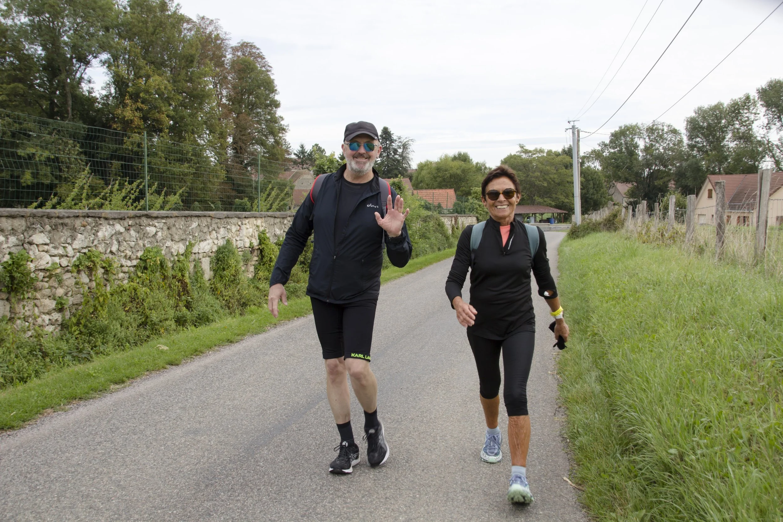 Deux personnes en tenue de course à pied marchent ou courent sur une route de campagne, elles portent des sunglasses et sourient, l'une des personnes fait un signe de la main.