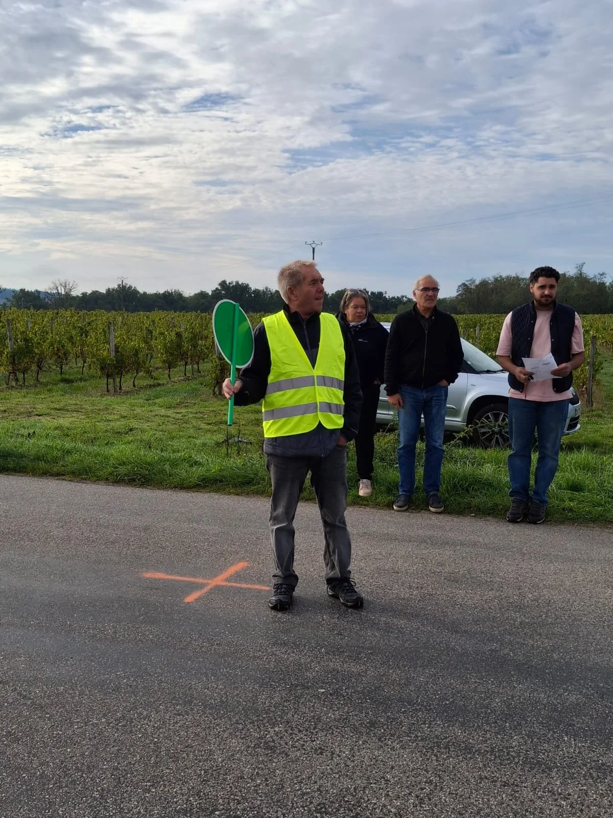 Un groupe de personnes, dont une portant un gilet réfléchissant jaune, est sur le bord d'une route à la campagne, avec un champ de vignes en arrière-plan, un ciel nuageux et une voiture derrière eux.