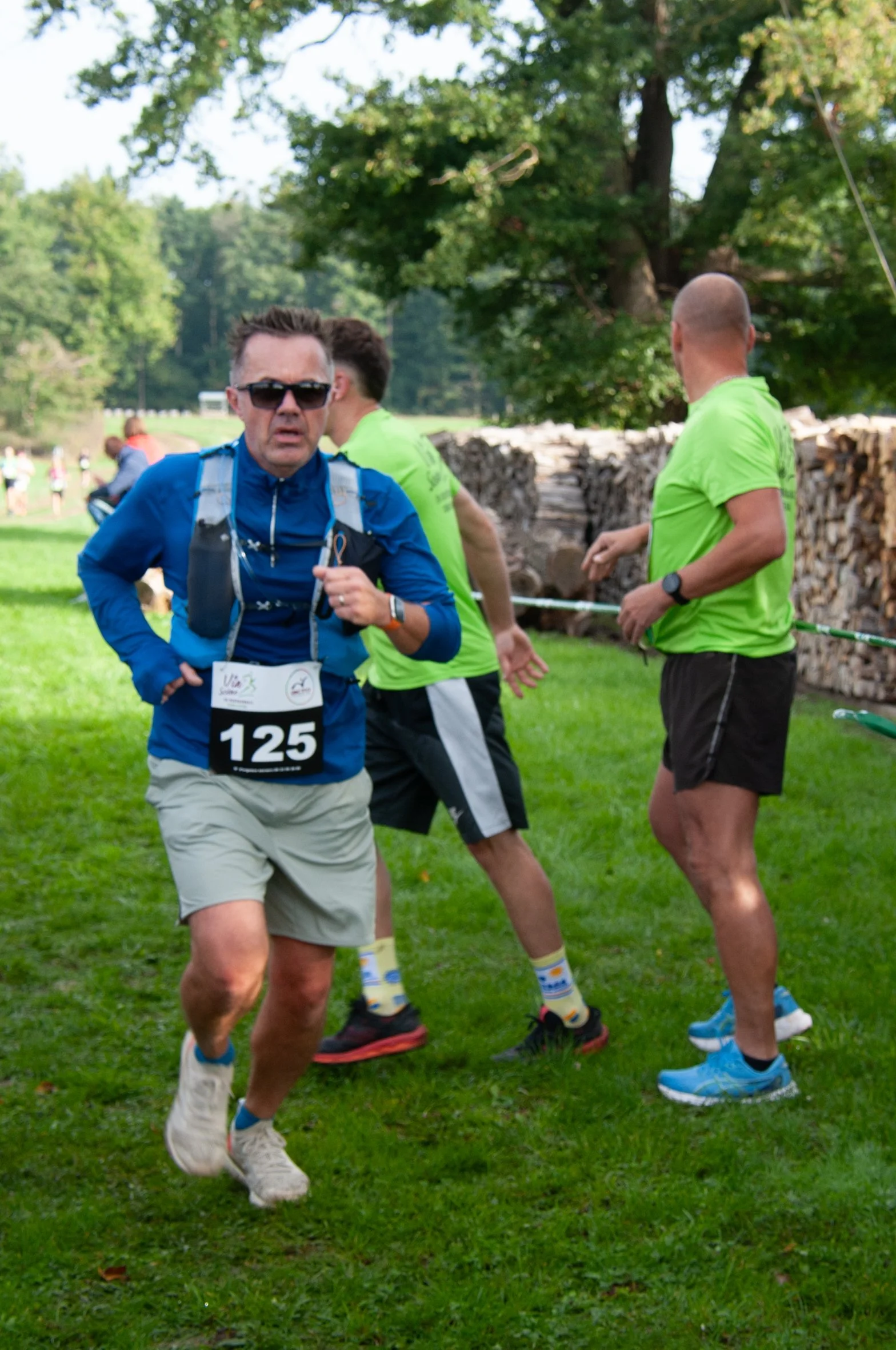 Un homme en tenue de course portant un dossard avec le numéro 125 court dans un parc lors d'une compétition, accompagné de deux autres coureurs en t-shirts verts, avec un fond d'arbres et de bûches de bois.
