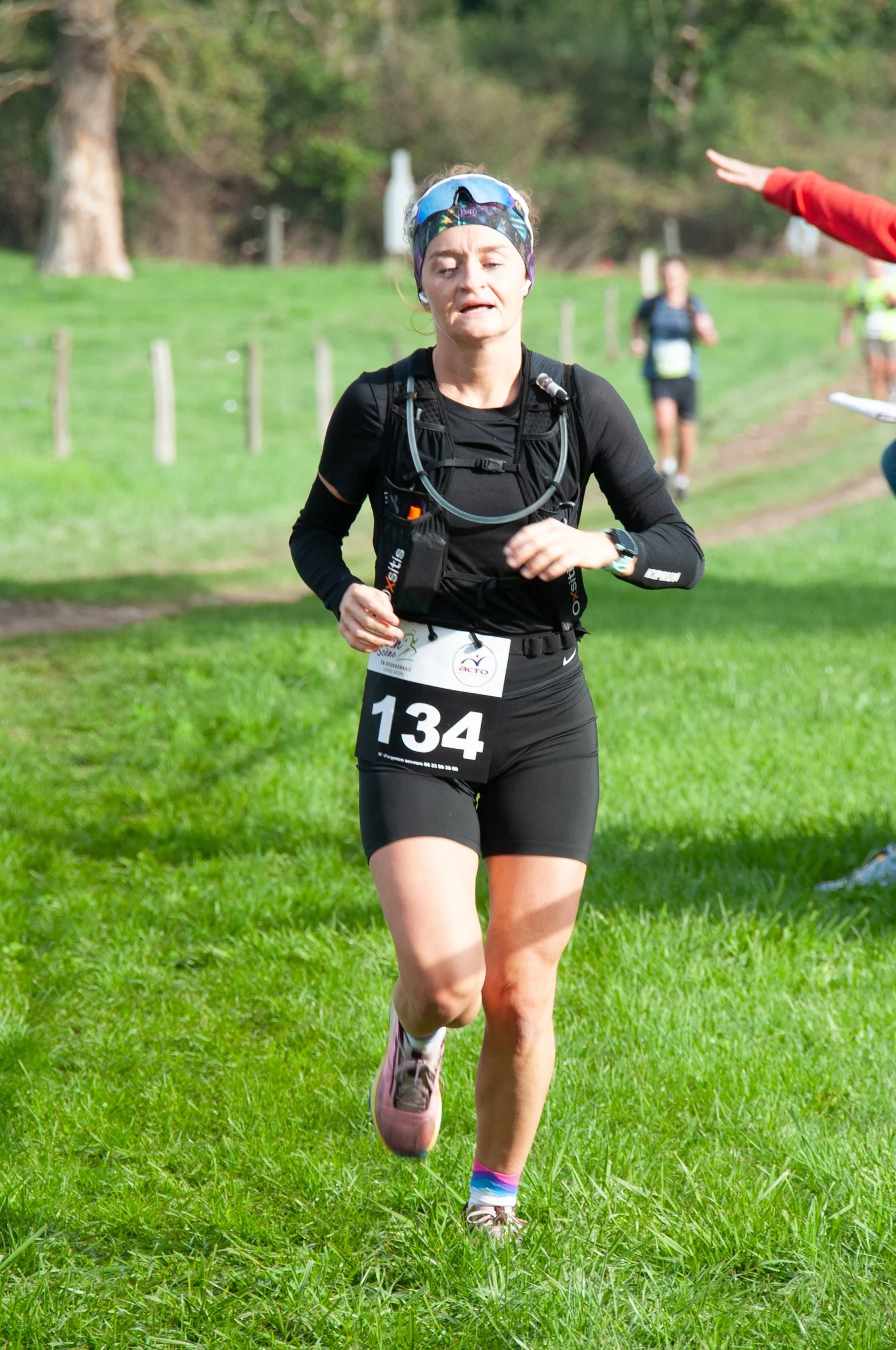 Une femme courait lors d'une course de trail en plein air, portant un numéro 134, une tenue de course noire, un casque de sport, et une montre, avec un terrain verdoyant en arrière-plan.
