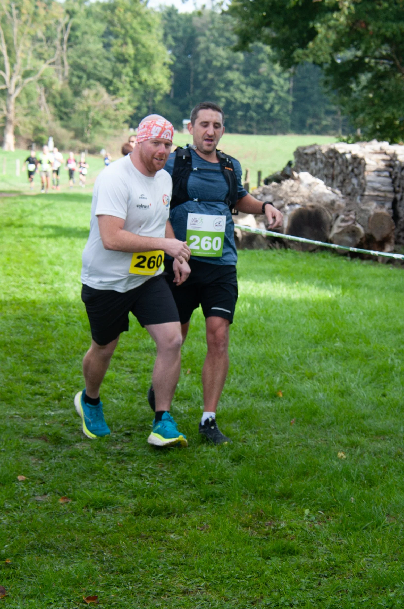 Deux hommes courent lors d'une course en extérieur dans une zone herbeuse, portant des numéros de course 260, avec un groupe de coureurs en arrière-plan et un environnement verdoyant.