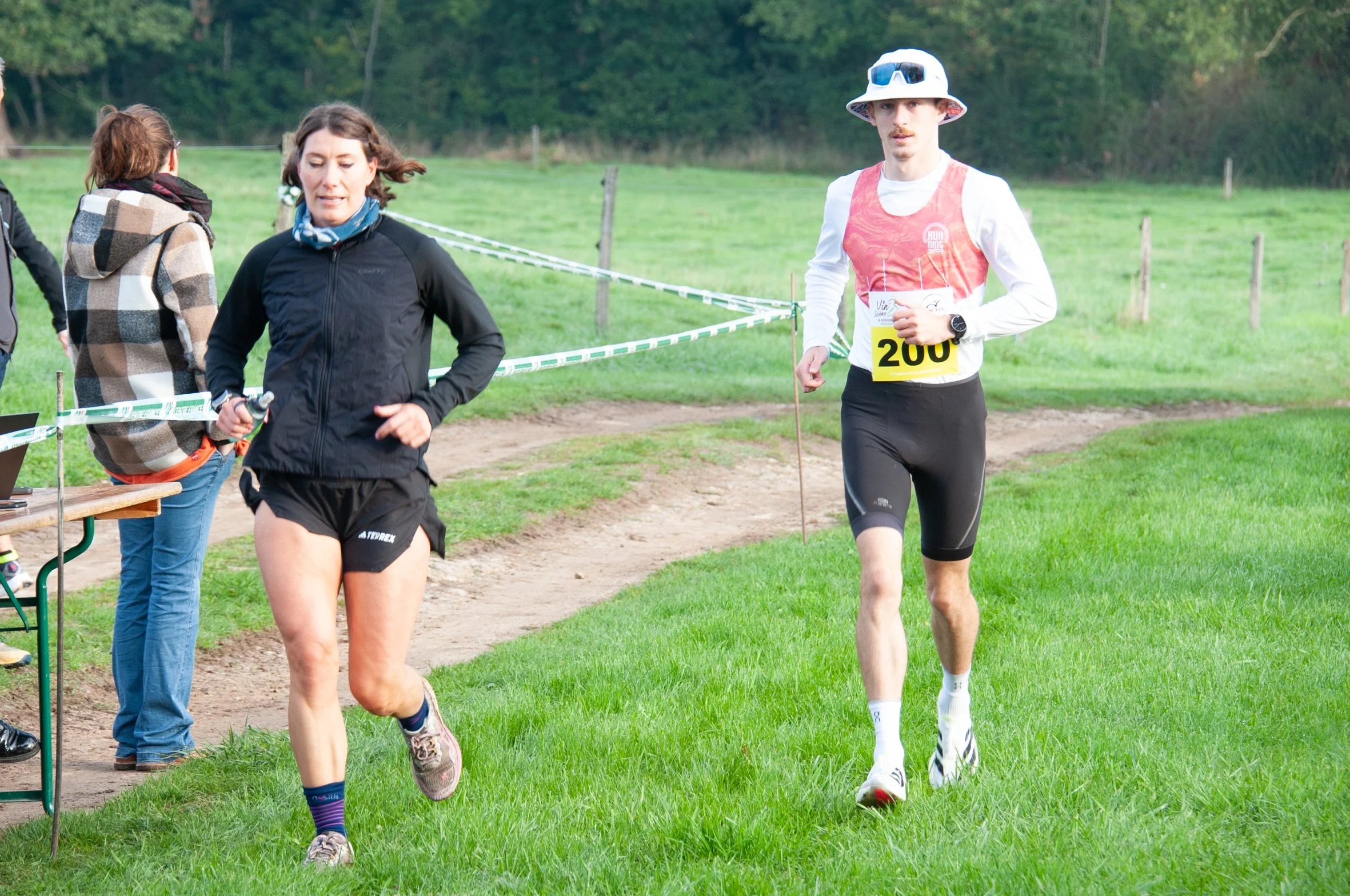 Deux coureurs, un homme et une femme, participent à une course en plein air. L'homme porte un débardeur rouge, un chapeau blanc, des lunettes de soleil, et un numéro 200. La femme porte une veste noire, un short noir, et court sur l'herbe. Il y a un 