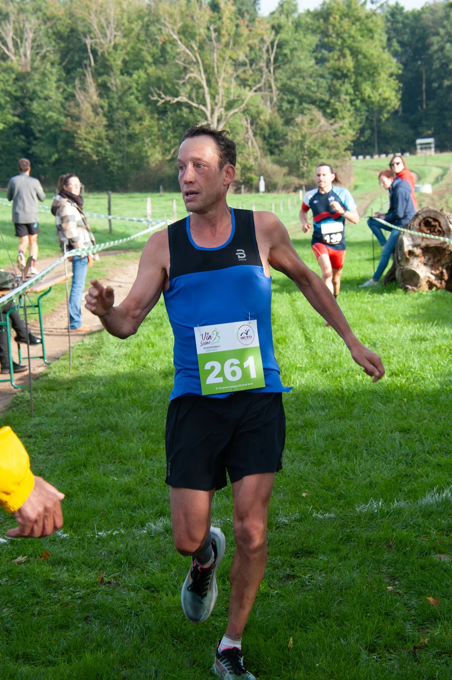 Un homme court lors d'une course en extérieur, portant un dossard numéro 261, dans un environnement verdoyant avec des autres coureurs et spectateurs en arrière-plan.