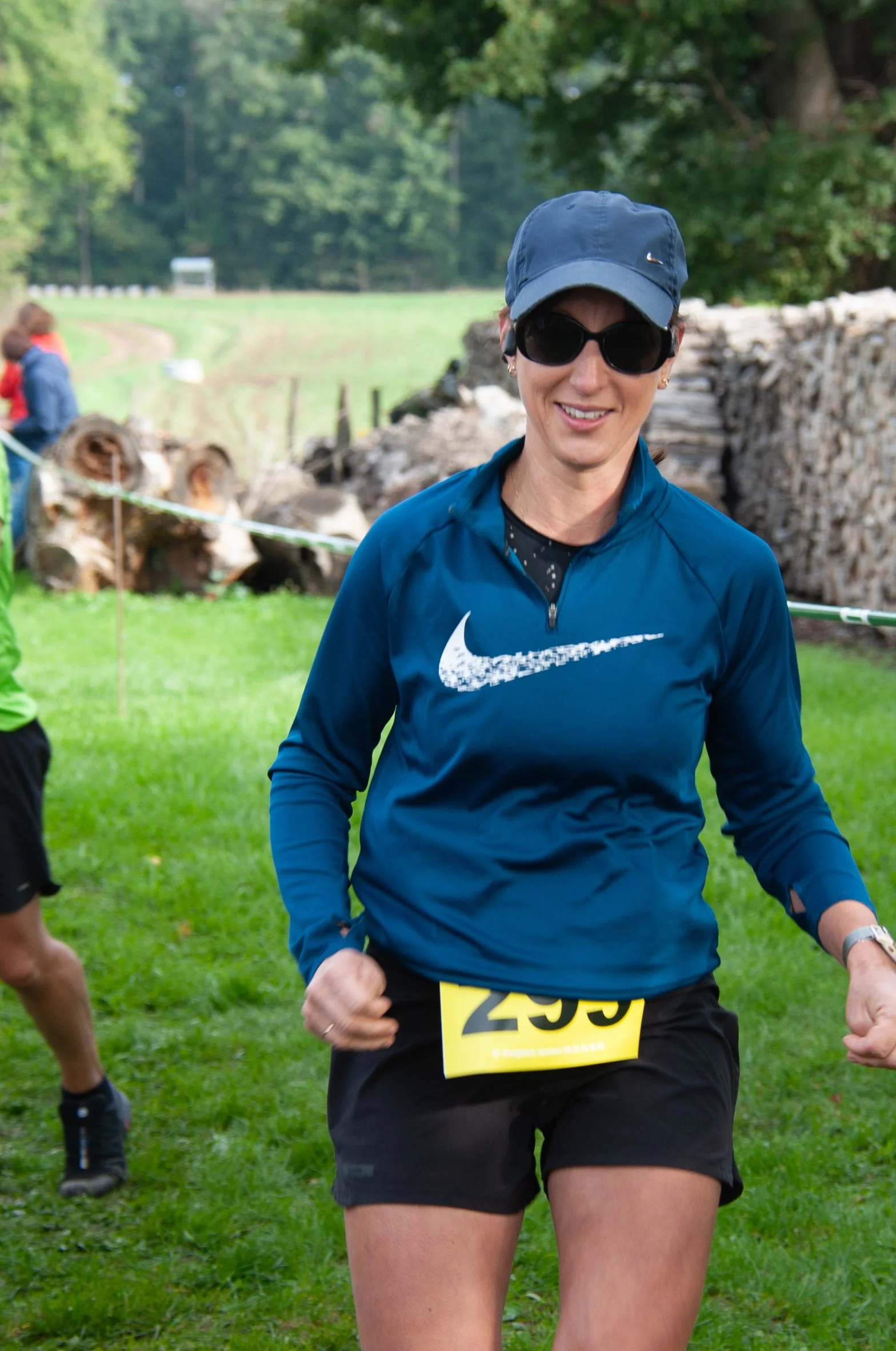 Une femme faisant une course, portant une veste bleue Nike, un chapeau bleu et des lunettes de soleil, en train de courir dans un parc verdoyant.