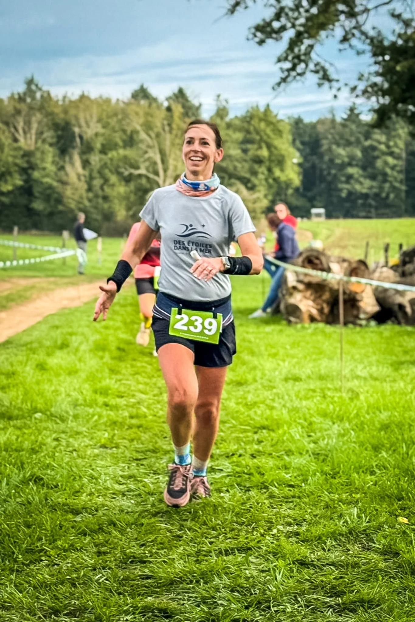 Une femme souriante court sur un chemin herbeux lors d'une course, portant un dossard avec le numéro 239, vêtue d'un t-shirt gris, short noir, et des accessoires de course, entourée d'un paysage verdoyant.