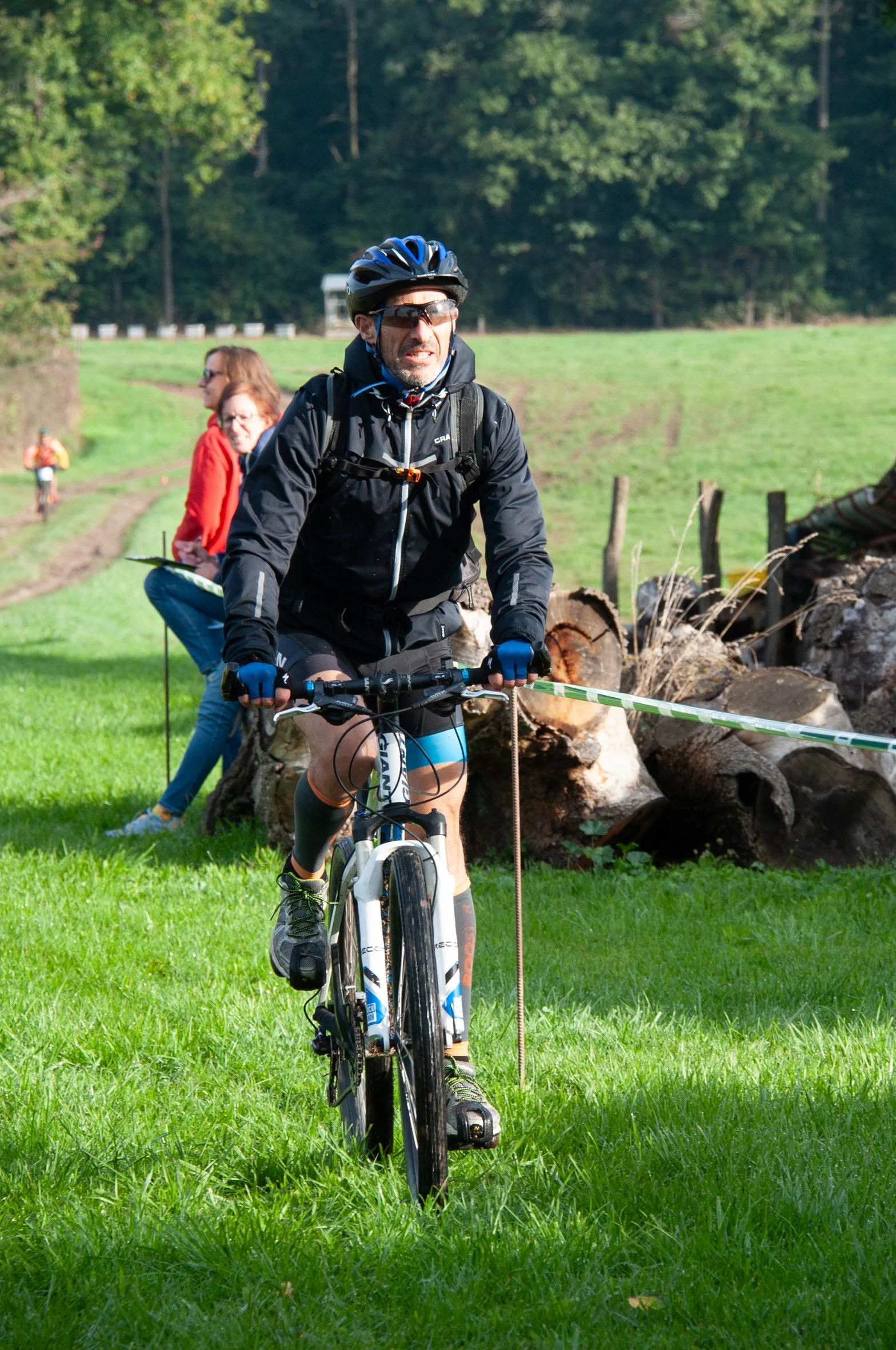Homme en VTT avec casque et lunettes, portant un sac à dos, roulant sur une pelouse verte lors d'une activité en plein air.