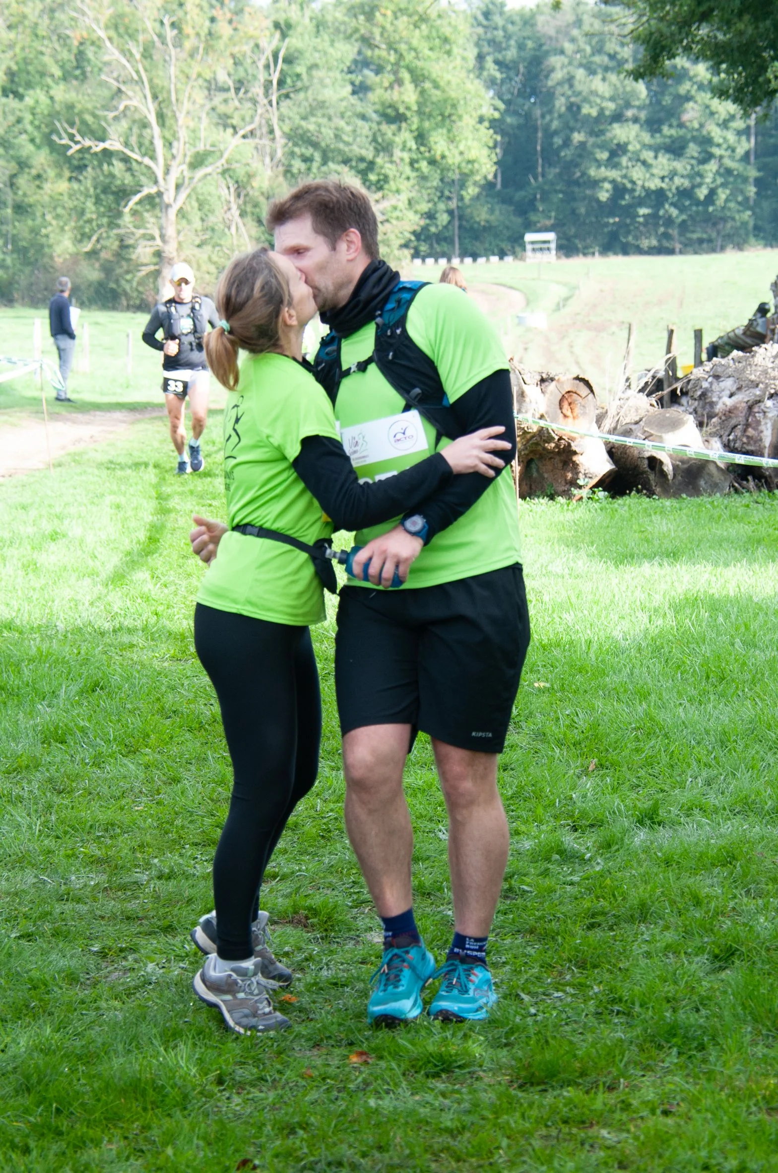 Deux coureurs en tenue de sport vert et noir échangent un baiser après une course en plein air, avec d'autres participants en arrière-plan.