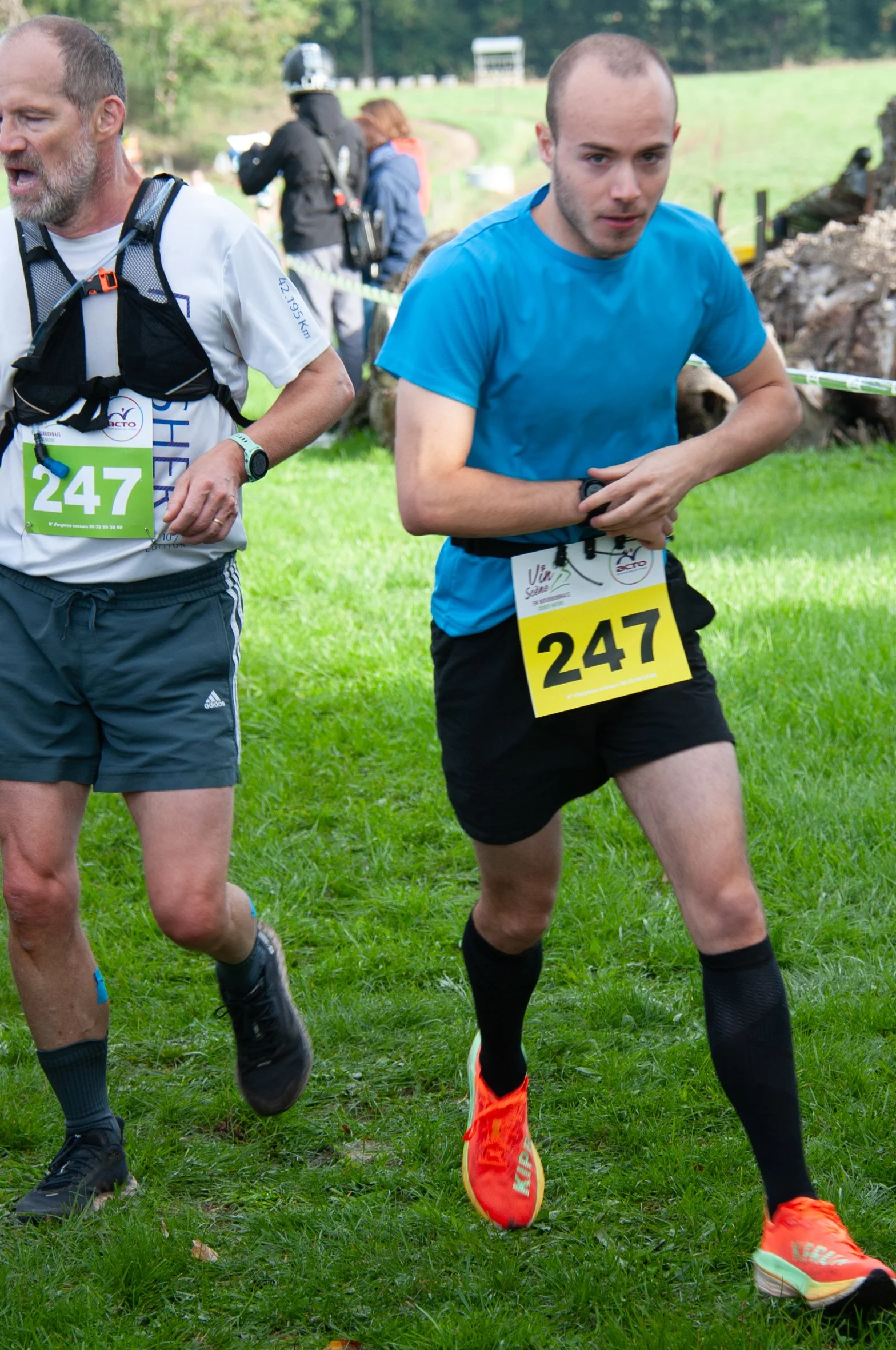 Deux coureurs participent à une course en plein air, portant un dossard avec le numéro 247, dans un environnement verdoyant.