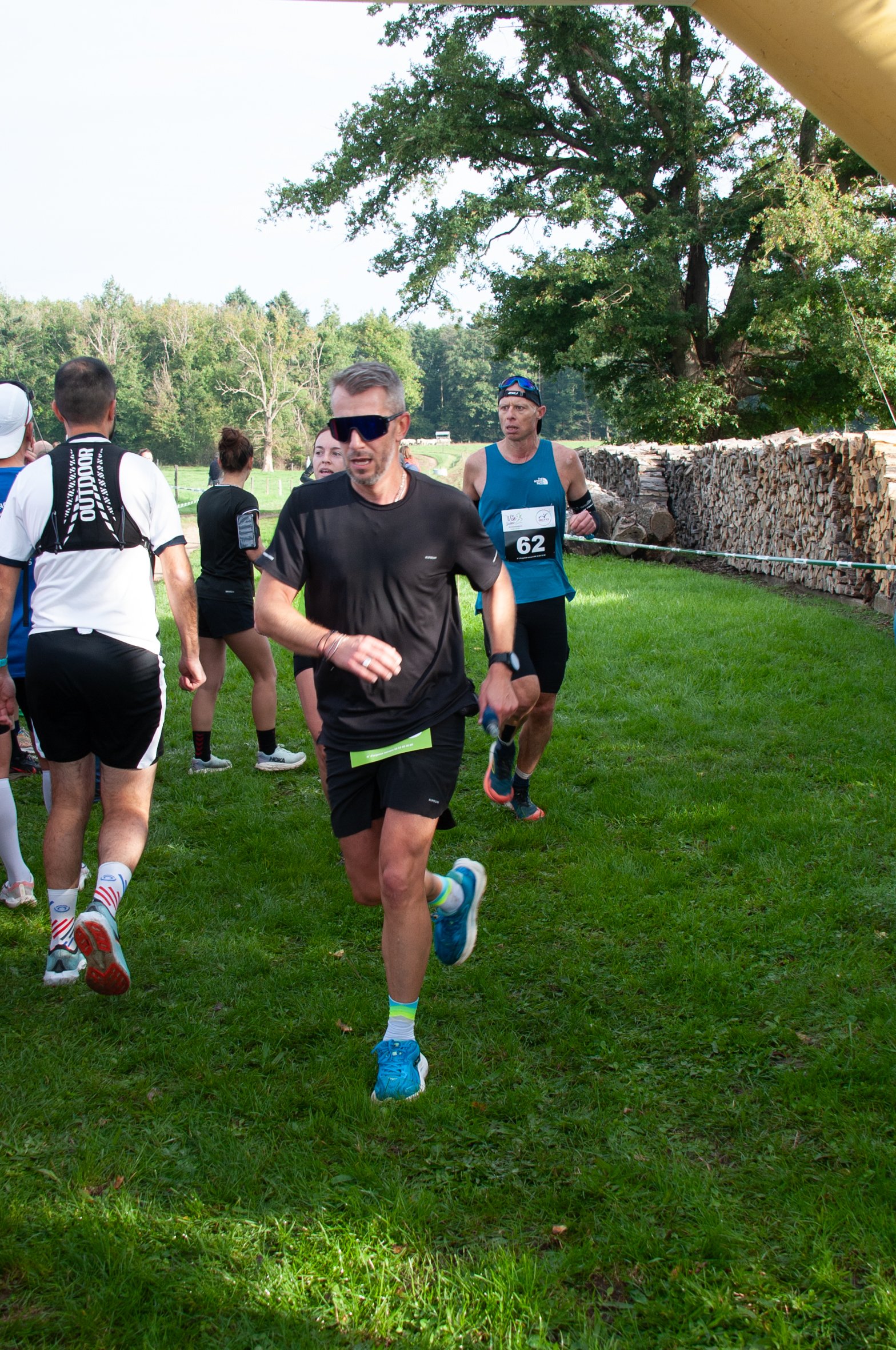 Des coureurs participent à une course en extérieur sur une pelouse verte, avec des arbres en arrière-plan et un vieux tas de bois le long du côté droit de l'image.