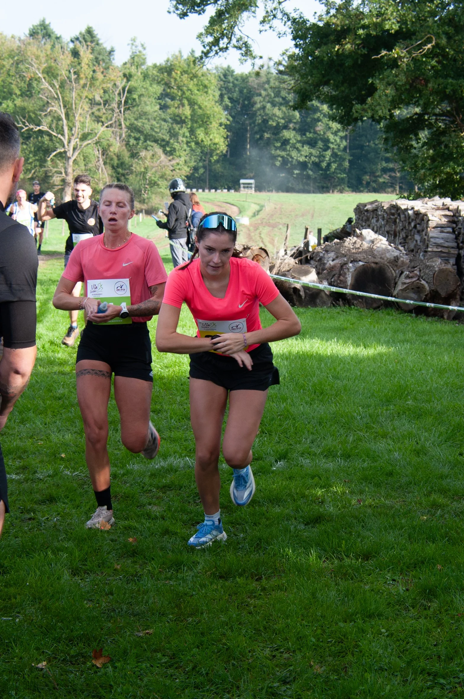 Deux femmes courent en dehors d'une course en utilisant des vêtements de sport de couleur rose, avec un environnement de parc ou de forêt en arrière-plan.