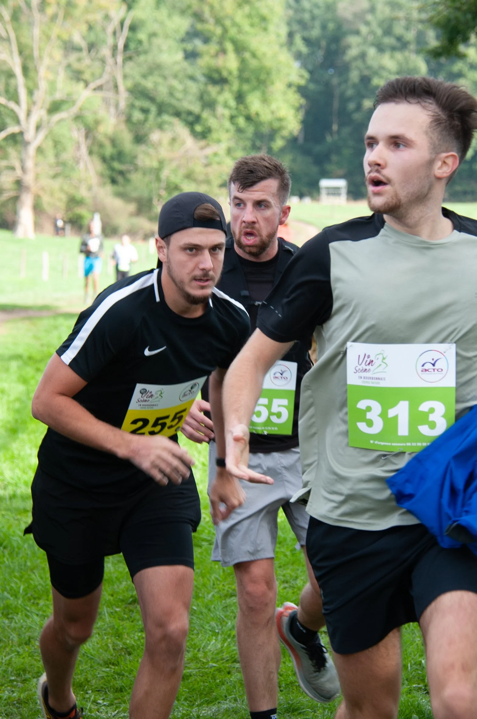 Trois hommes courent lors d'une course en extérieur, portant des numéros de dossard, dans un parc avec des arbres en arrière-plan.