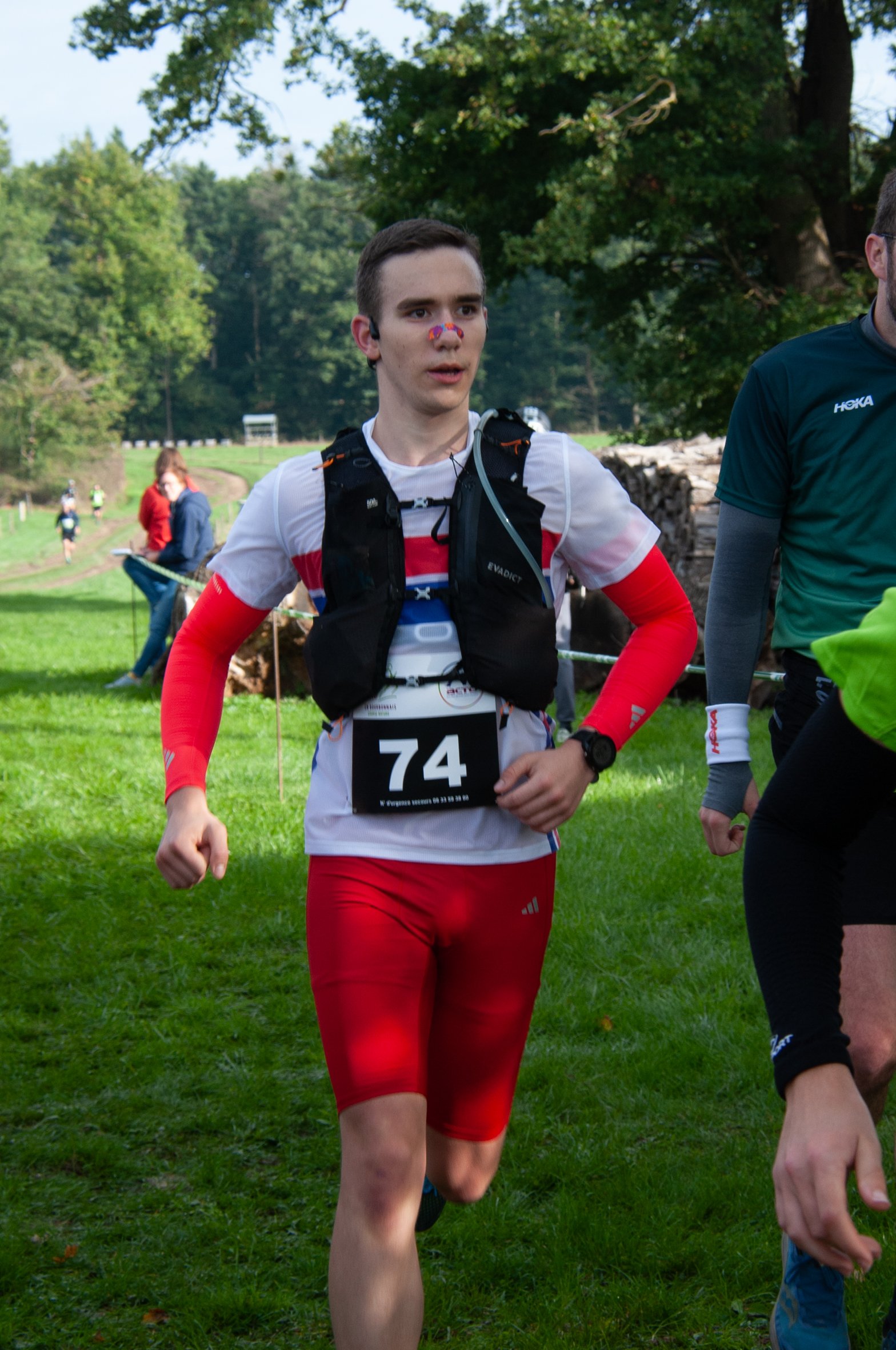Jeune homme courant lors d'une course en plein air, portant un dossard numéro 74, une tenue de sport rouge et blanche, un podomètre, et un gilet d'hydratation, avec un arrière-plan de paysage verdoyant.