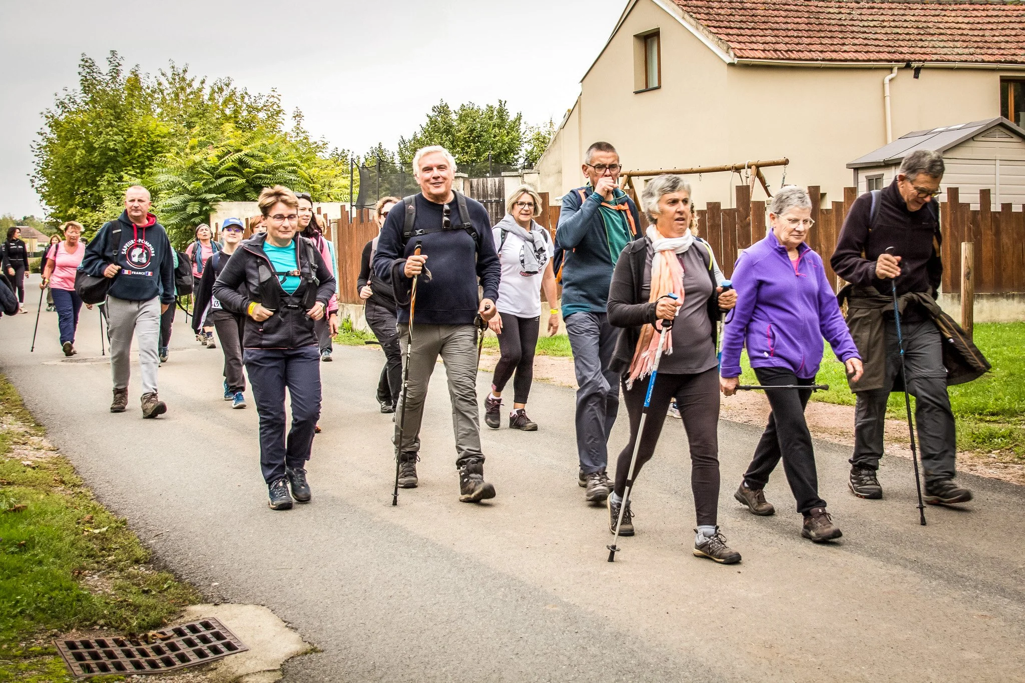 Groupe de personnes âgées faisant une marche en plein air sur un chemin de campagne, avec des maisons et des arbres en arrière-plan.