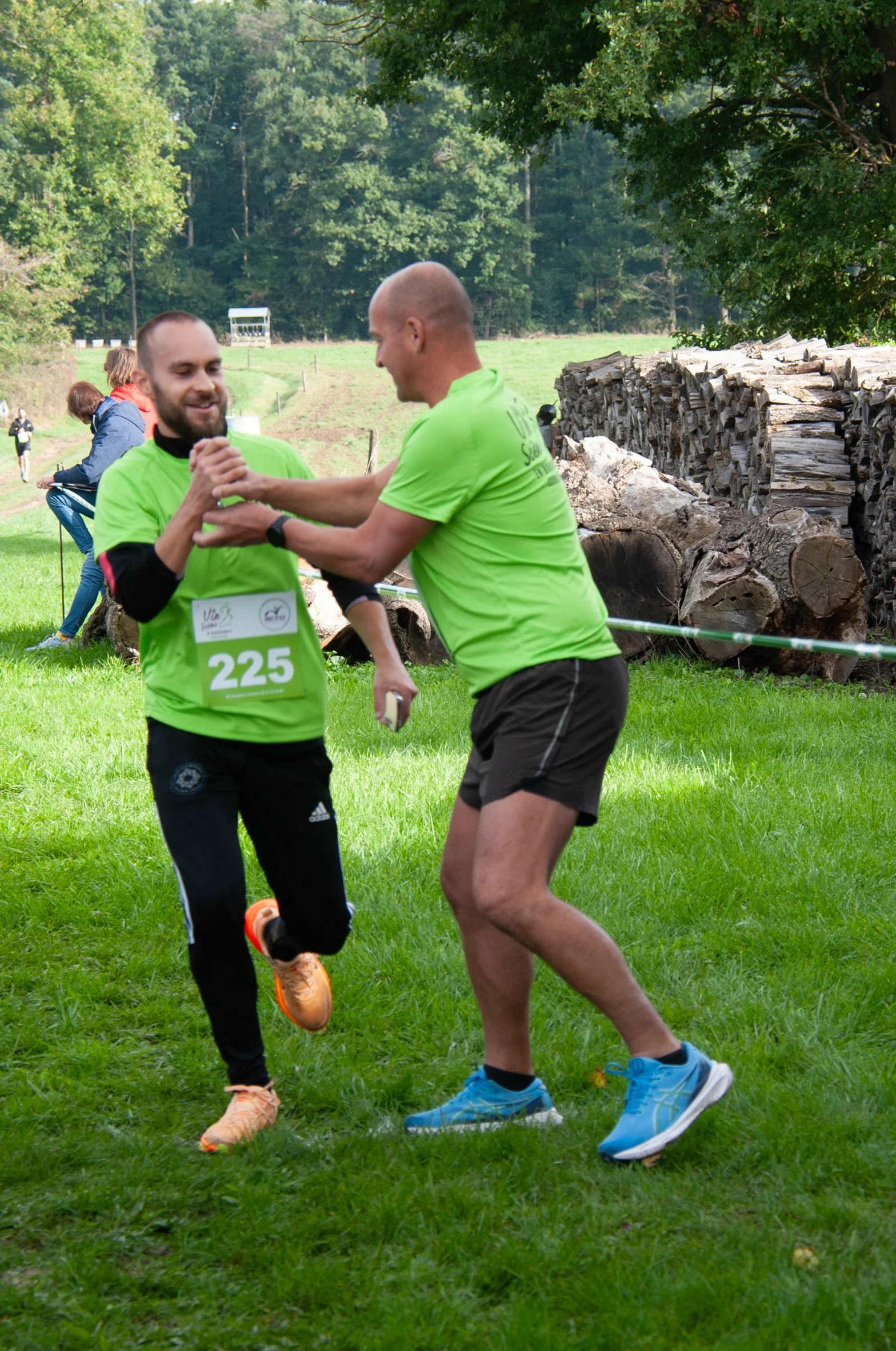 Deux hommes en vêtements de course se félicitent et se donnent la main dans un parc vert lors d'une course, avec d'autres participants en arrière-plan, sous un arbre, à côté de tas de bois.