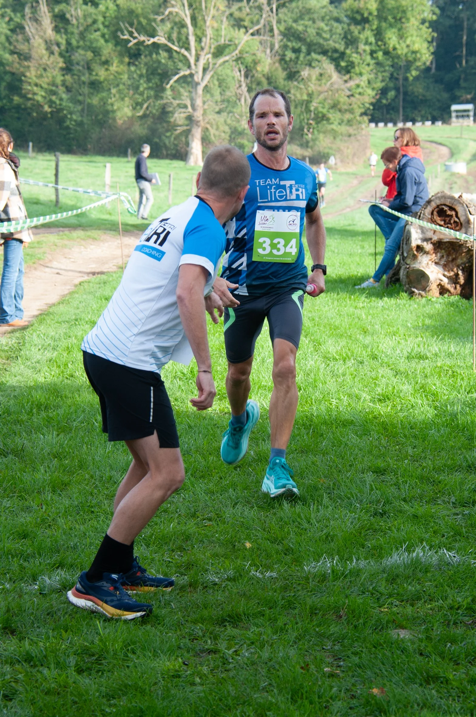 Deux hommes courent lors d'une compétition de course en plein air dans une zone verte boisée. Un homme porte un numéro 334 et un t-shirt bleu avec l'inscription 'TEAM Life Tri'. L'autre homme porte un t-shirt blanc avec des lignes bleues. Il semble l