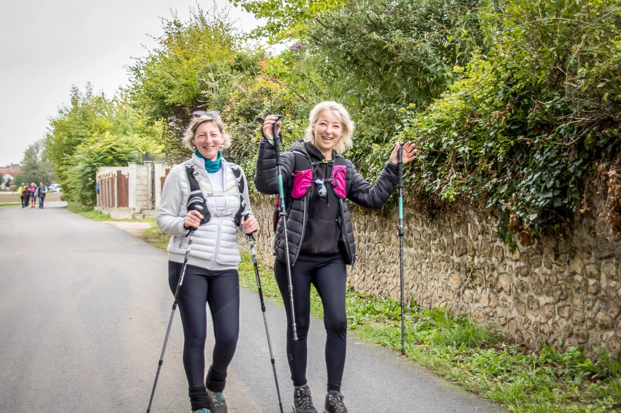 Deux femmes souriantes font de la randonnée avec des bâtons de marche sur un chemin bordé de verdure.