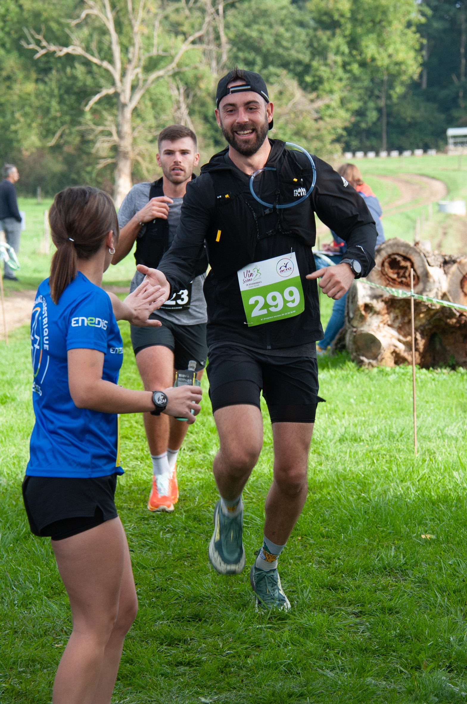 Un homme souriant, portant un dossard numéro 299, participe à une course en plein air, et échange un salut avec une bénévole. Deux autres coureurs le suivent derrière, dans un environnement naturel verdoyant.