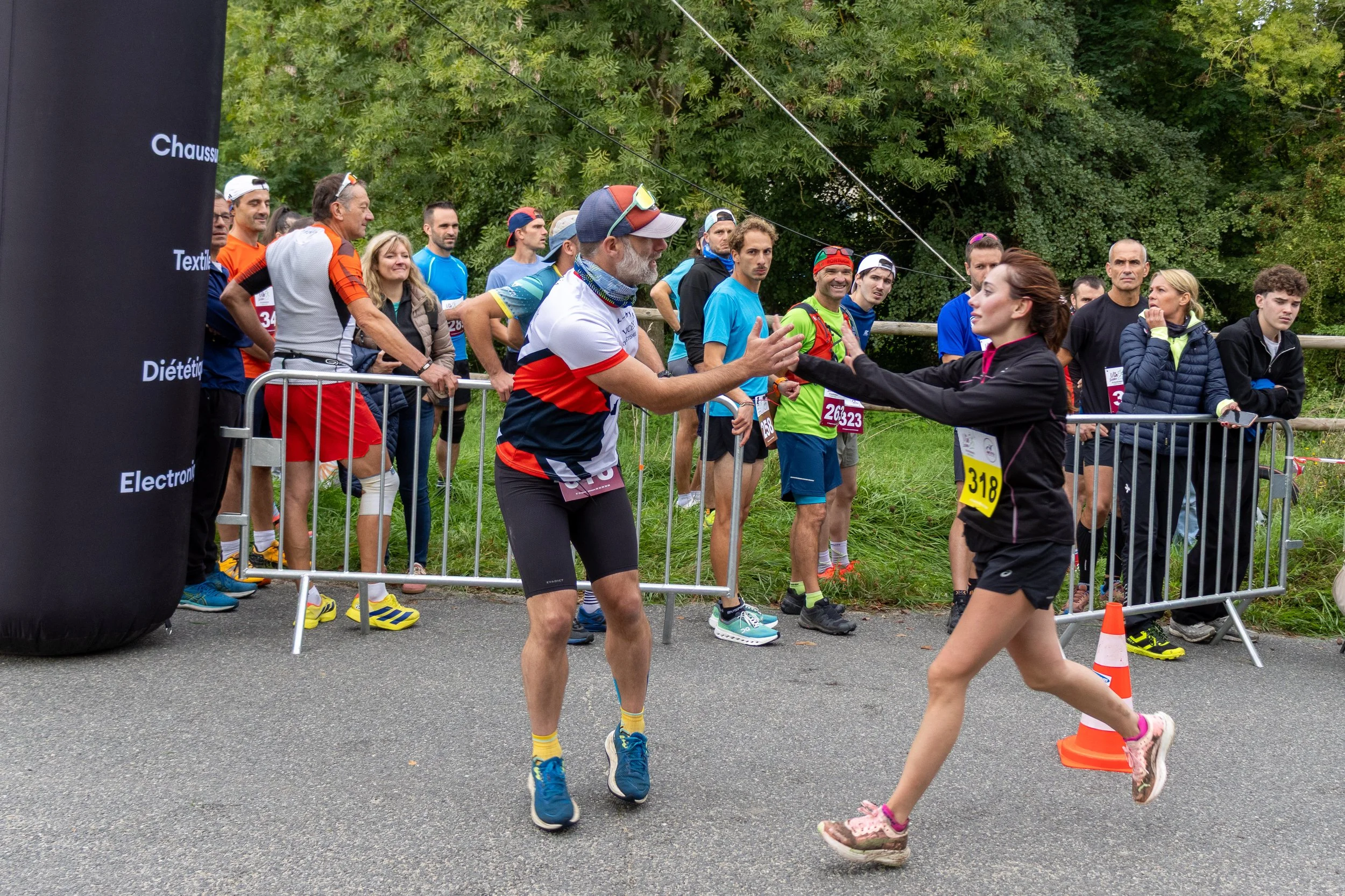 Un groupe de coureurs au début d'une course, l'un d'eux est en train de saluer ou de faire un geste de encouragement à une femme qui court vers lui, tous sont derrière une barrière de sécurité, avec des arbres en arrière-plan.