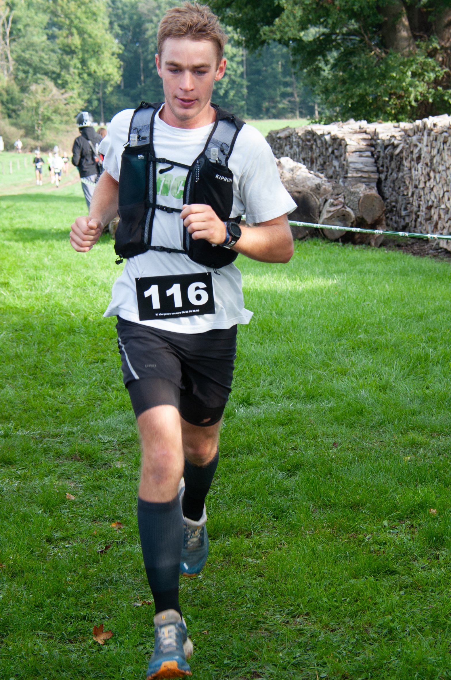 Un coureur participant à une course dans un parc verdoyant, portant un dossard numéro 116, une montre, un t-shirt blanc, un short noir et un sac à dos.